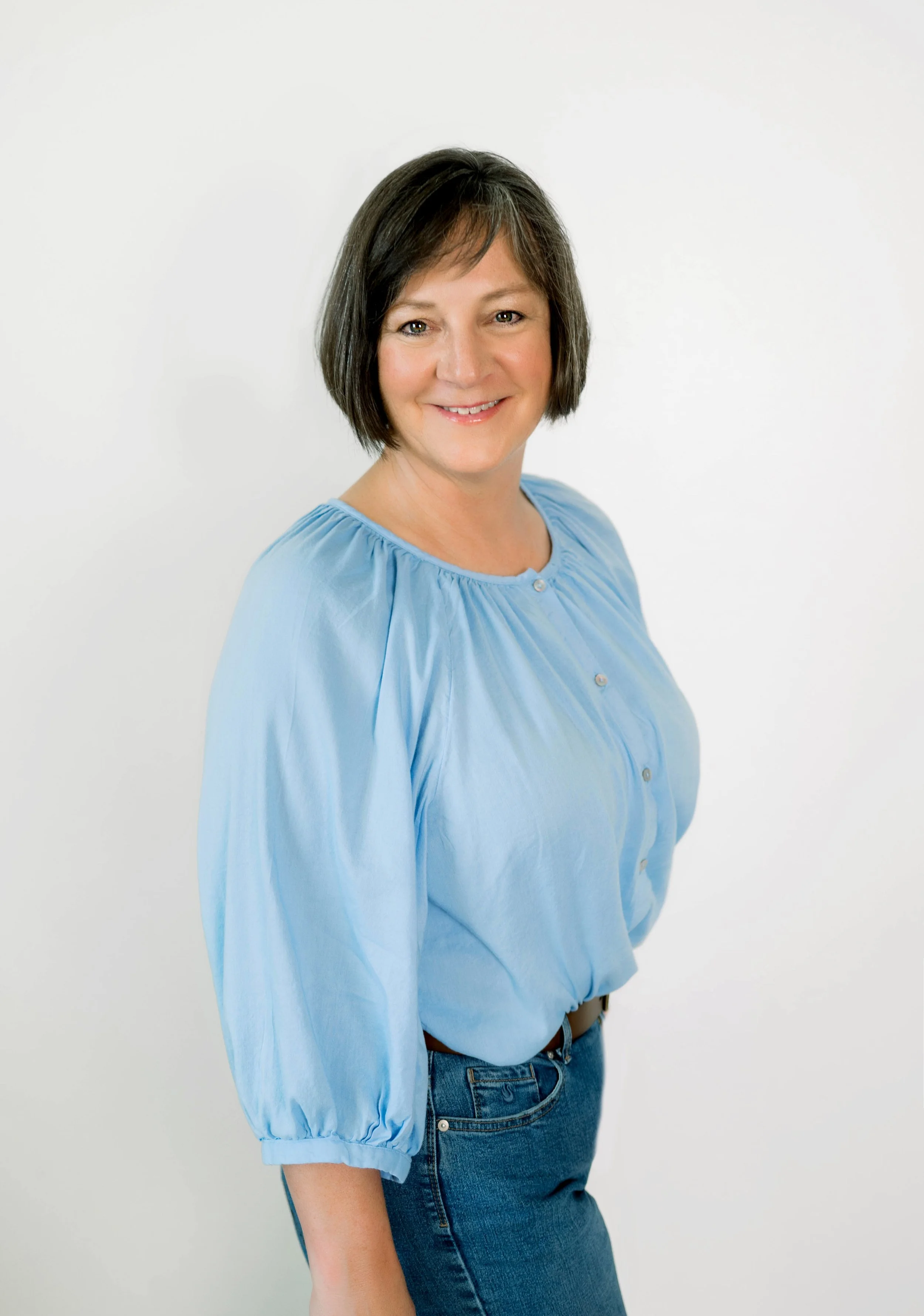 A woman with shoulder-length dark hair wearing a light blue blouse and blue jeans, smiling against a plain white background.