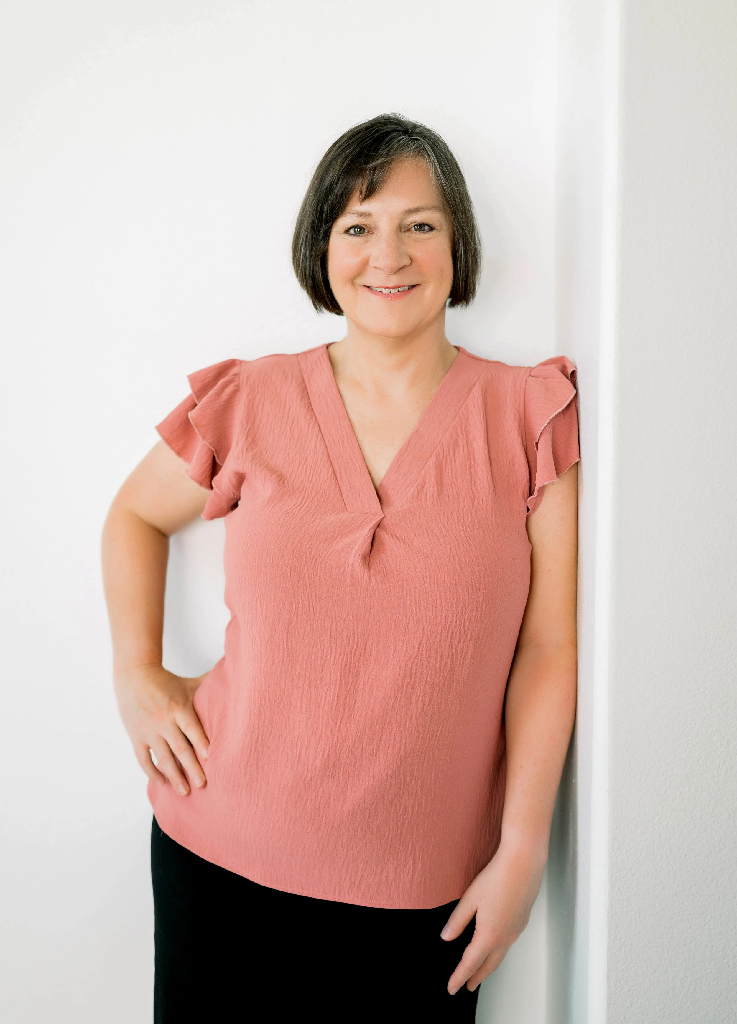 A woman with short dark hair wearing a pink top with ruffled sleeves, smiling and standing against a white wall.