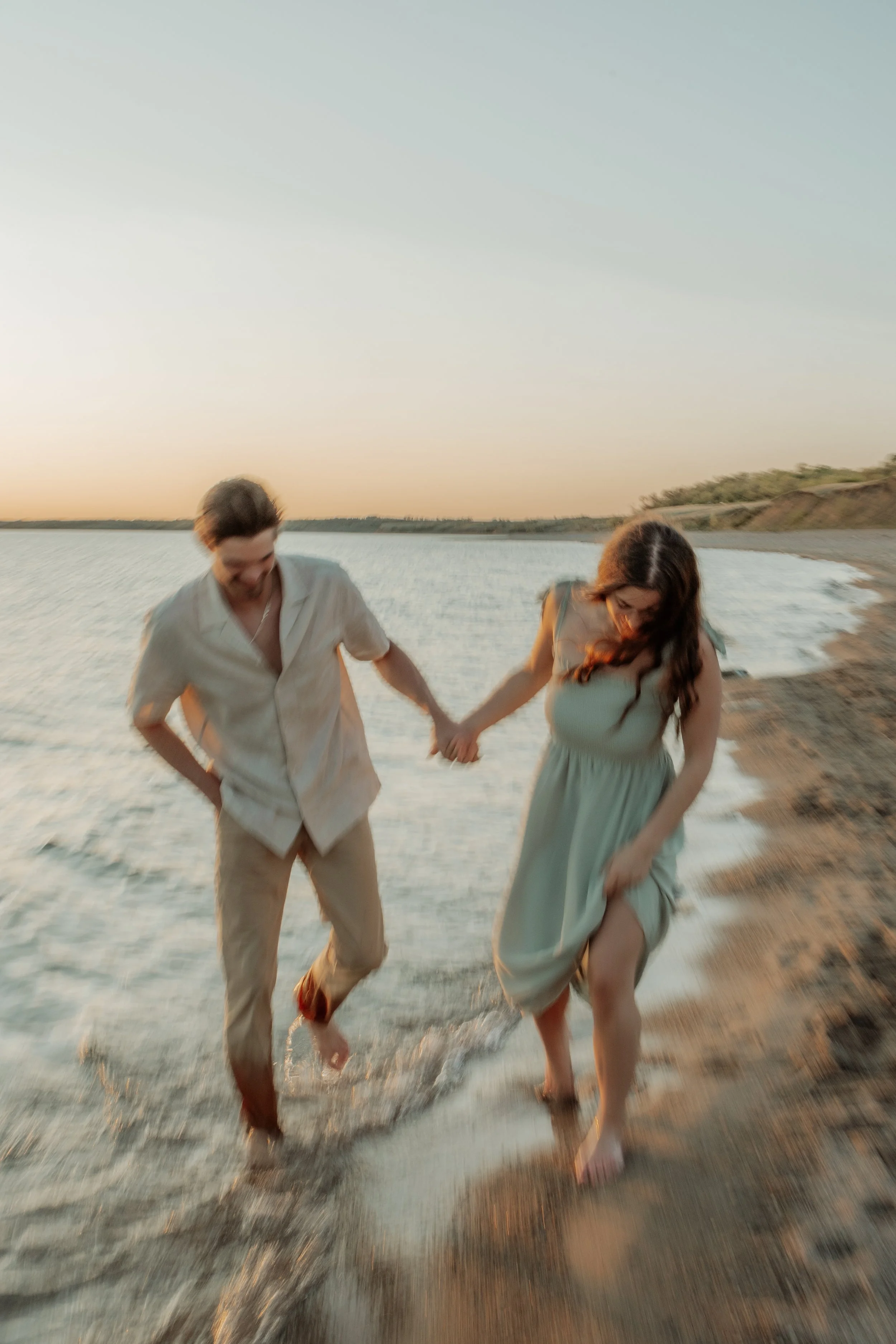 A couple holding hands and walking by the water on a beach at sunset.