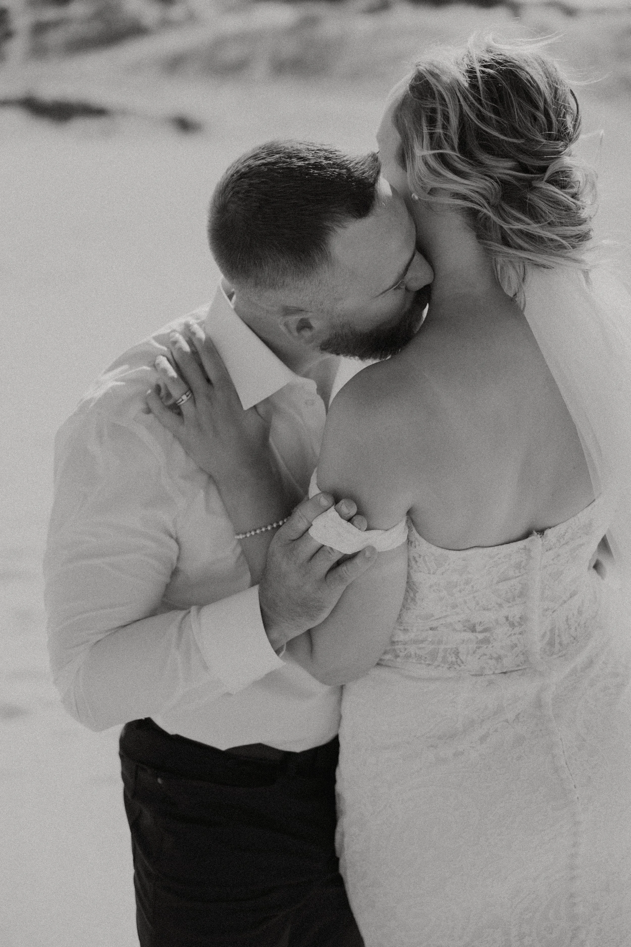 A black and white photo of a man and woman embracing, with the man kissing the woman's neck, on a beach.