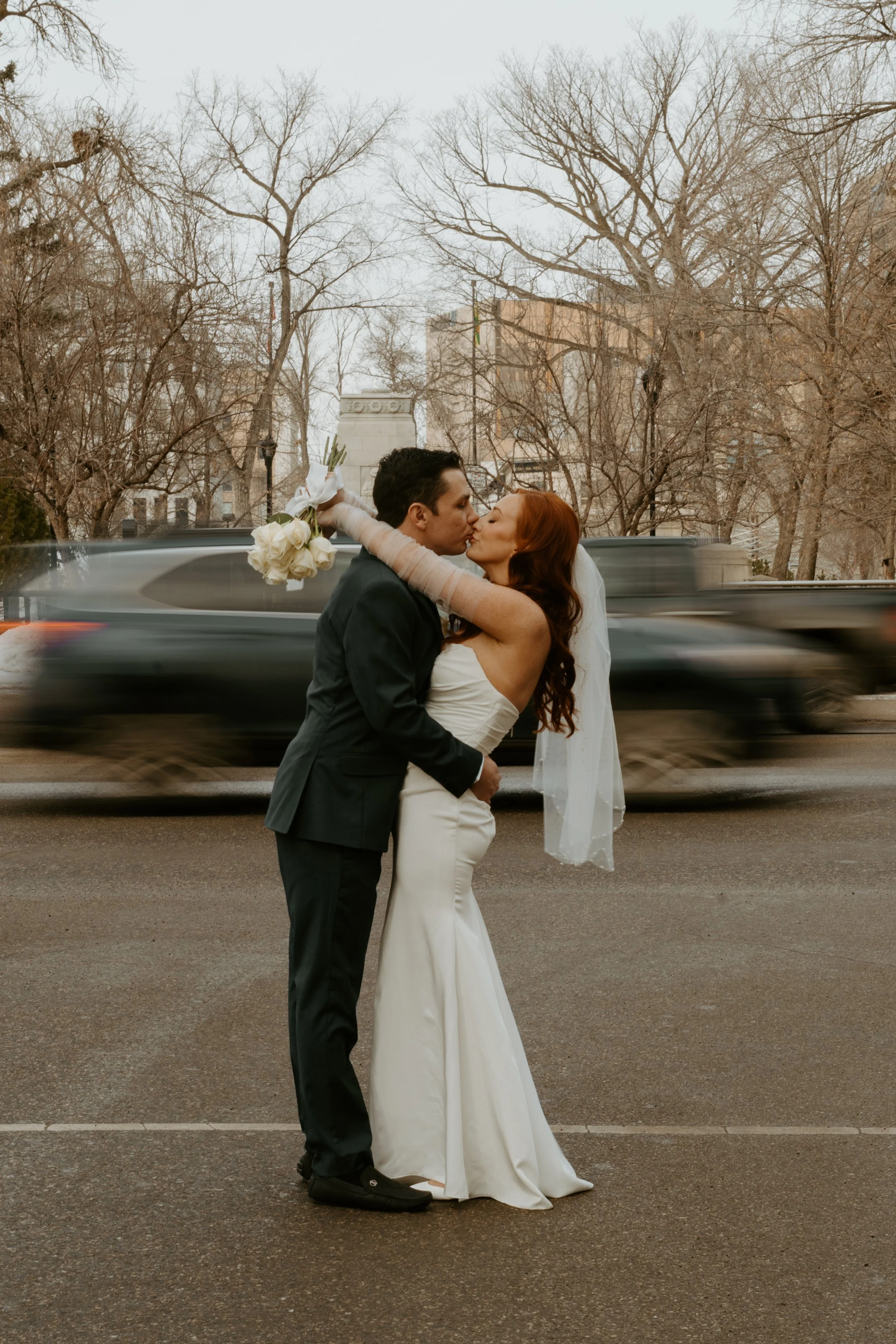 A bride and groom sharing a kiss on a city street during a wedding photoshoot. The bride is holding a bouquet of white roses and has long, wavy red hair. The groom is dressed in a black suit. A car is driving by behind them, appearing blurred due to 