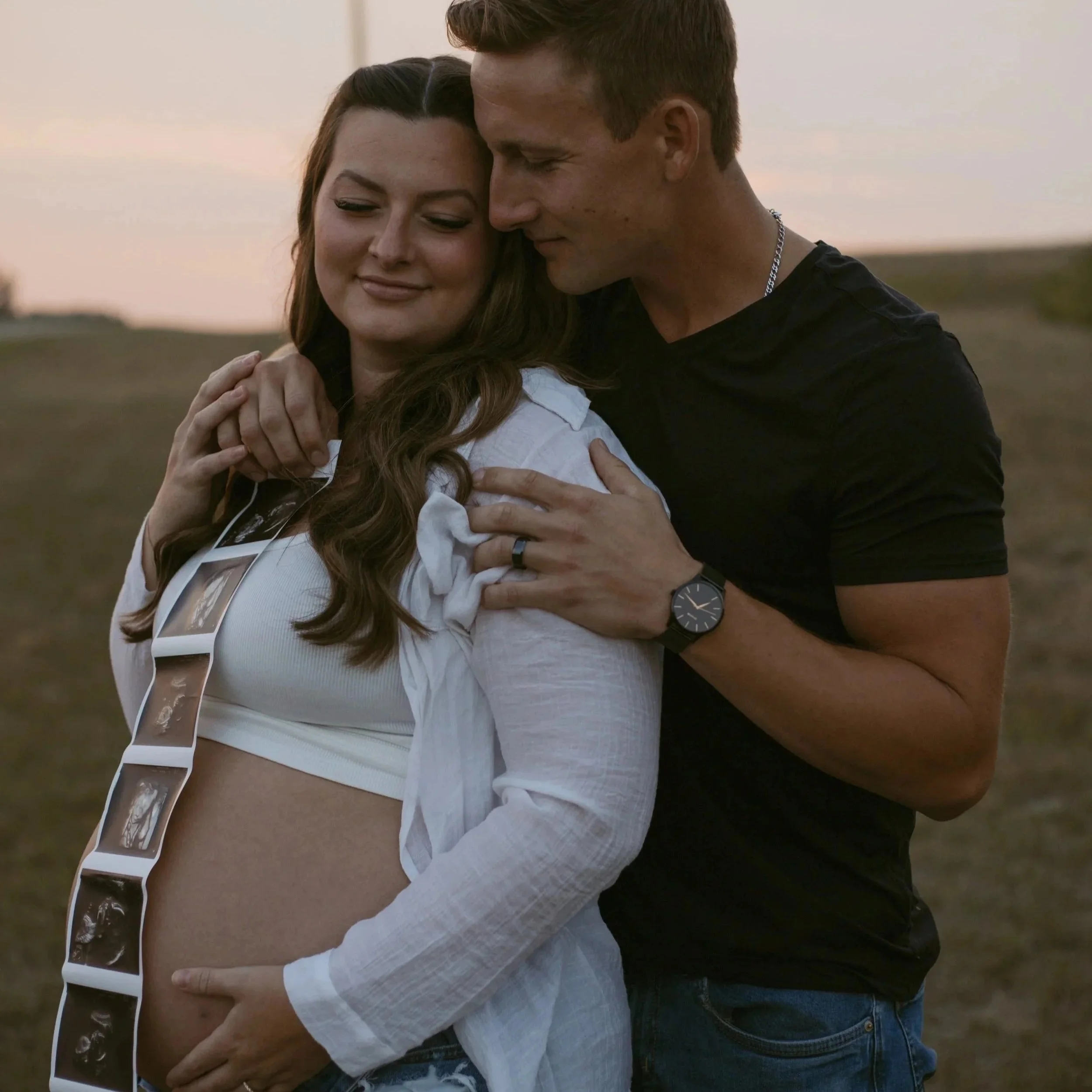 A couple sitting outdoors, leaning their foreheads together and smiling, surrounded by trees with autumn foliage.