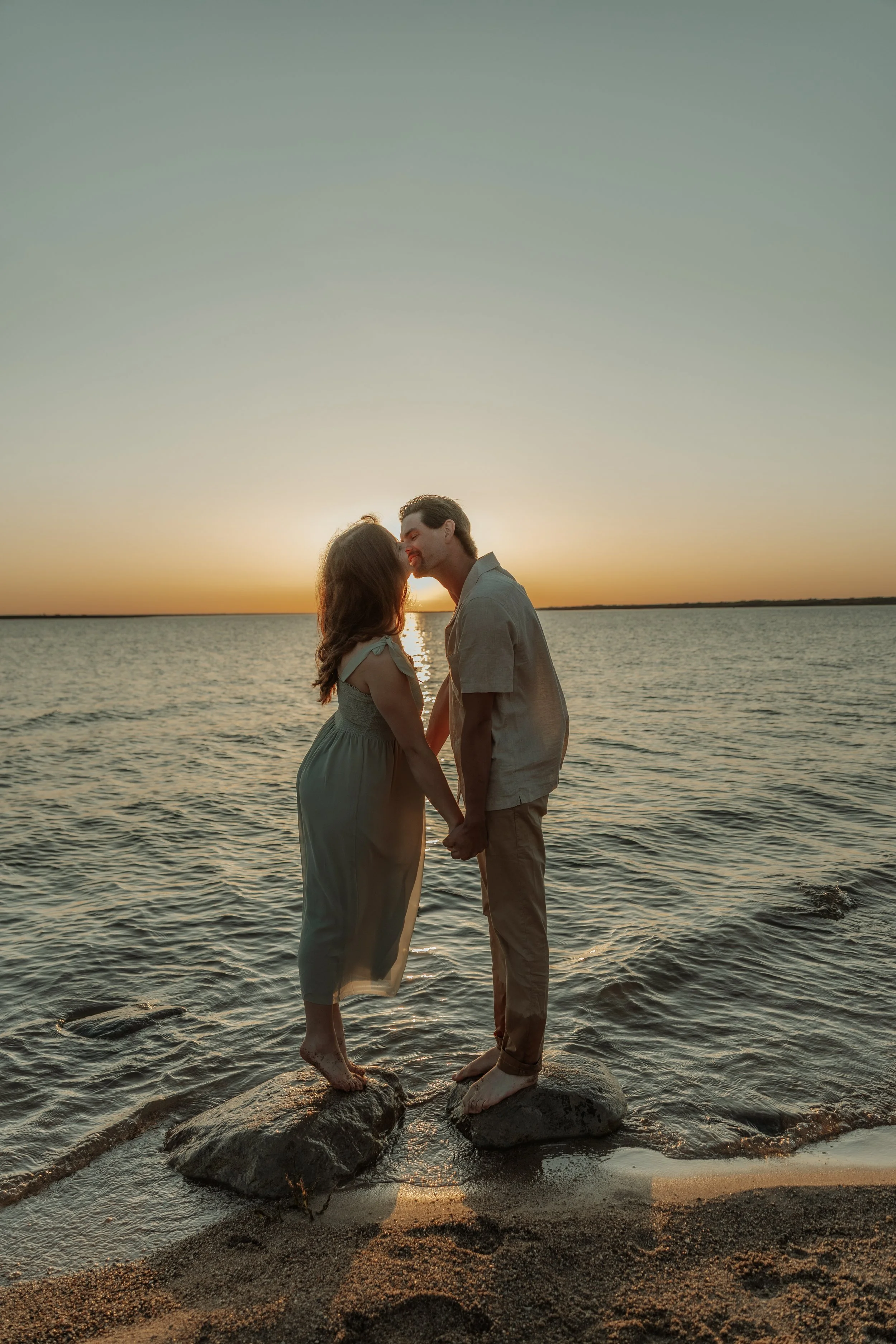 A couple standing on rocks in the water holding hands and touching foreheads during sunset.