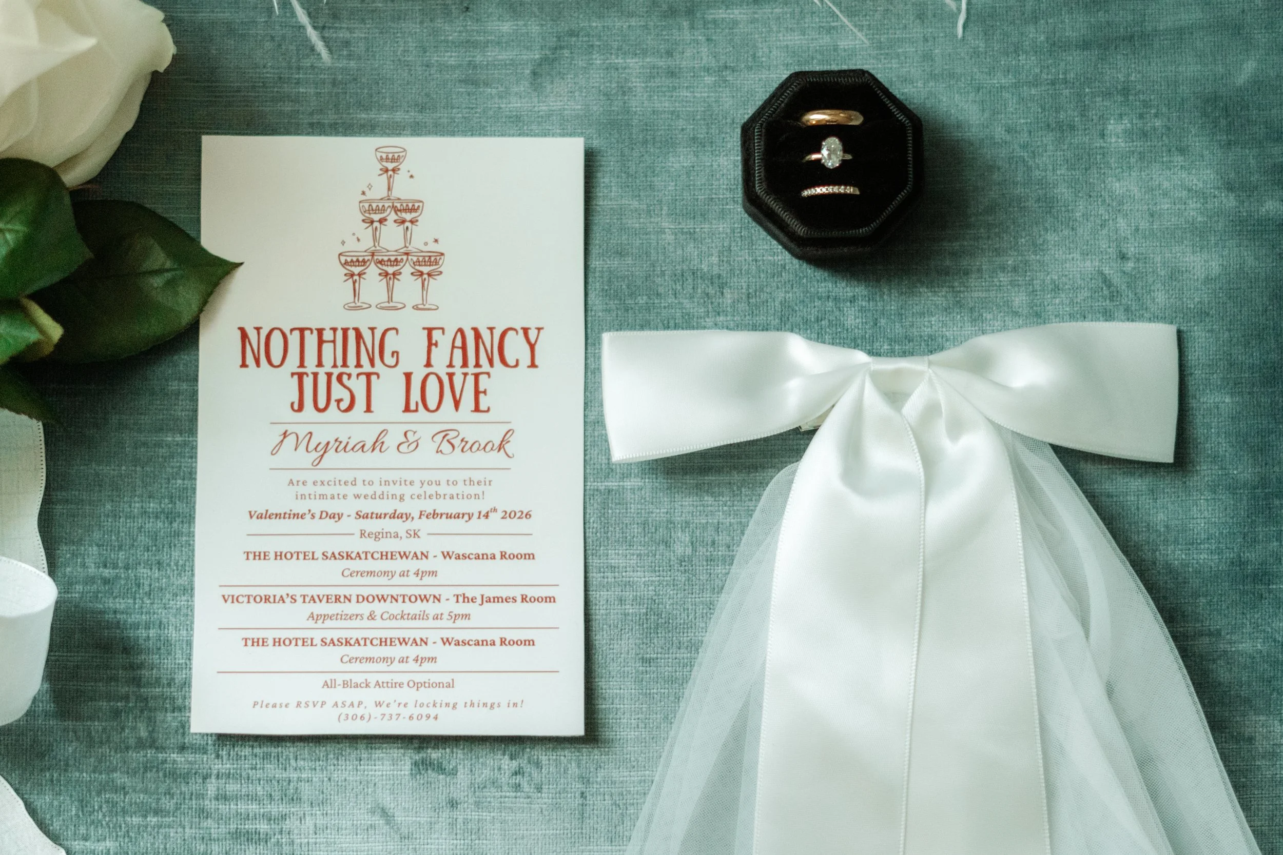 Wedding invitation for Myriah and Broak with a champagne glass illustration at the top, details about the wedding events at The Hotel Saskatchewan and Victoria's Tavern, with a white ribbon and a ring box with rings next to it, on a teal fabric surfa