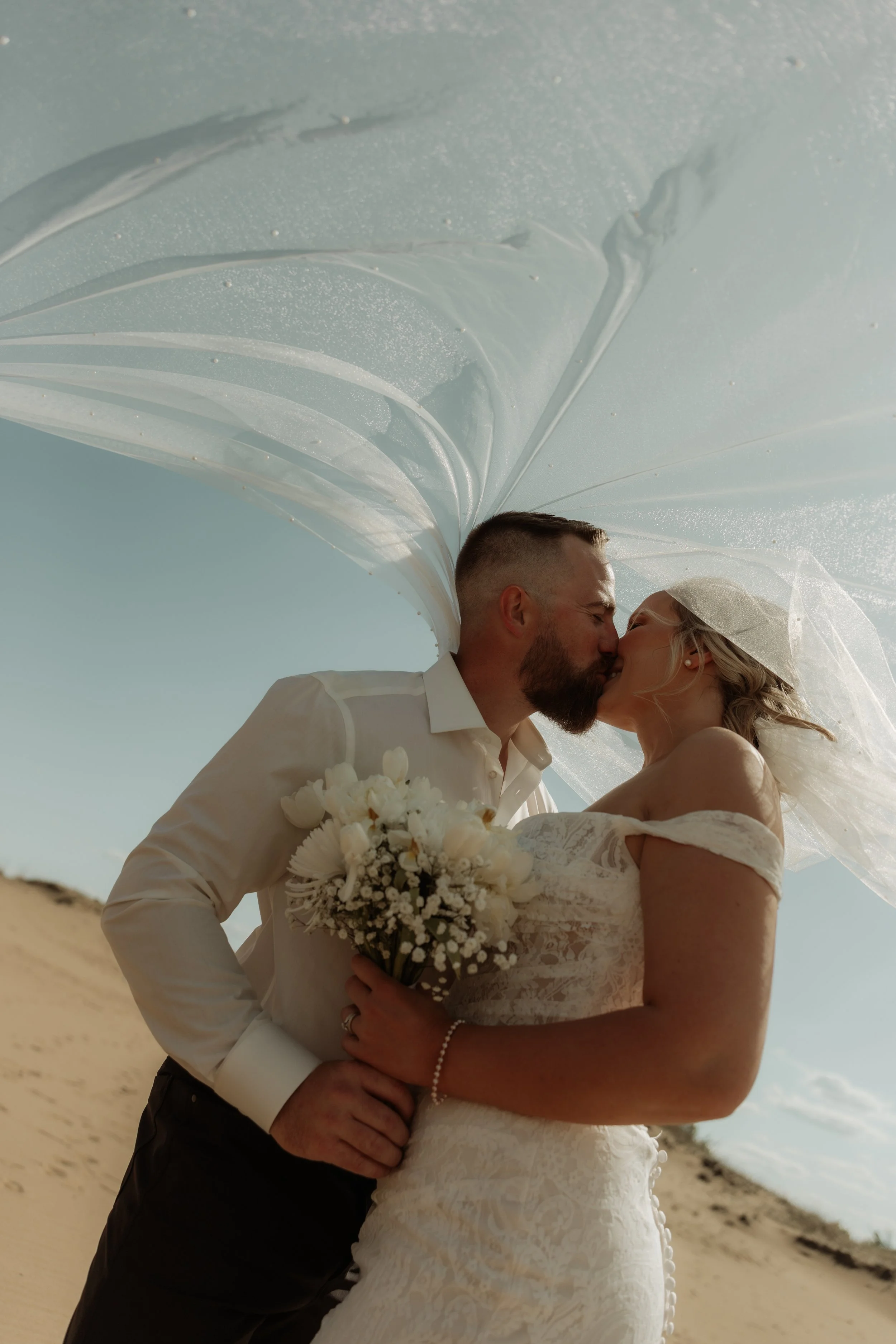 A bride and groom kiss under a wedding veil on a sandy outdoors beach with a blue sky.