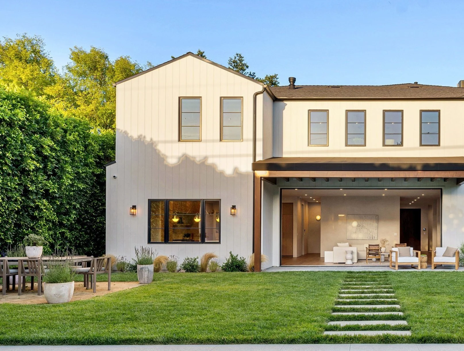 Modern two-story house with white exterior, large windows, and a covered patio. Green lawn and outdoor seating area with potted plants in the yard. Clear blue sky in the background.