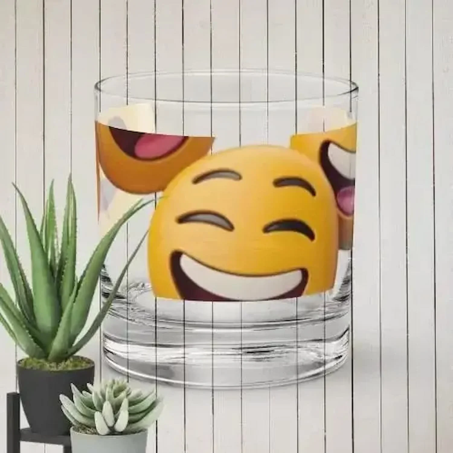 A glass of water with emojis of smiling and laughing faces on it, placed next to two potted succulent plants, with a white wooden background.