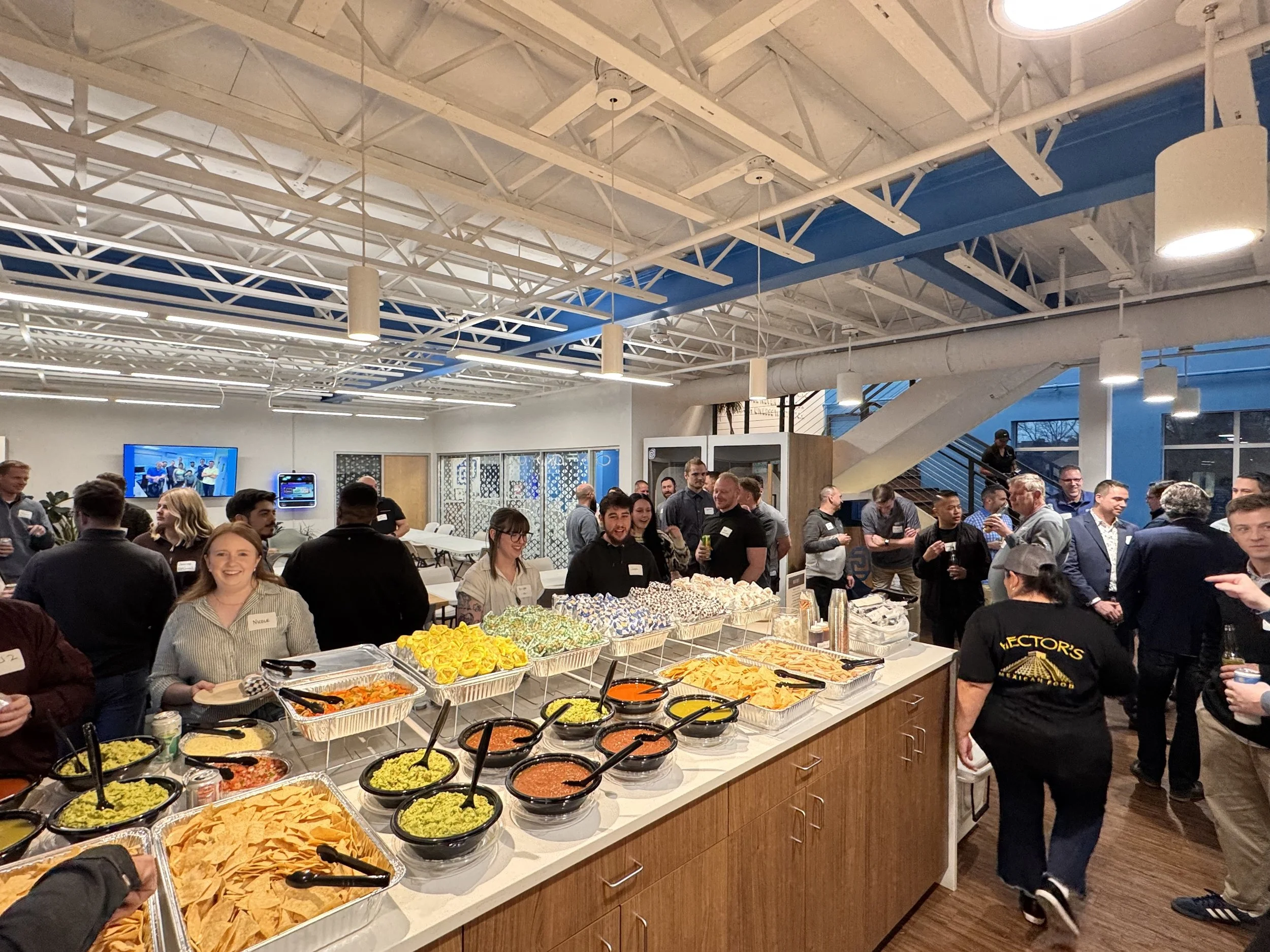 A buffet table with various dishes including guacamole, salsa, chips, and other Mexican foods set up at a social gathering. People are mingling and smiling in a large, well-lit, indoor space.
