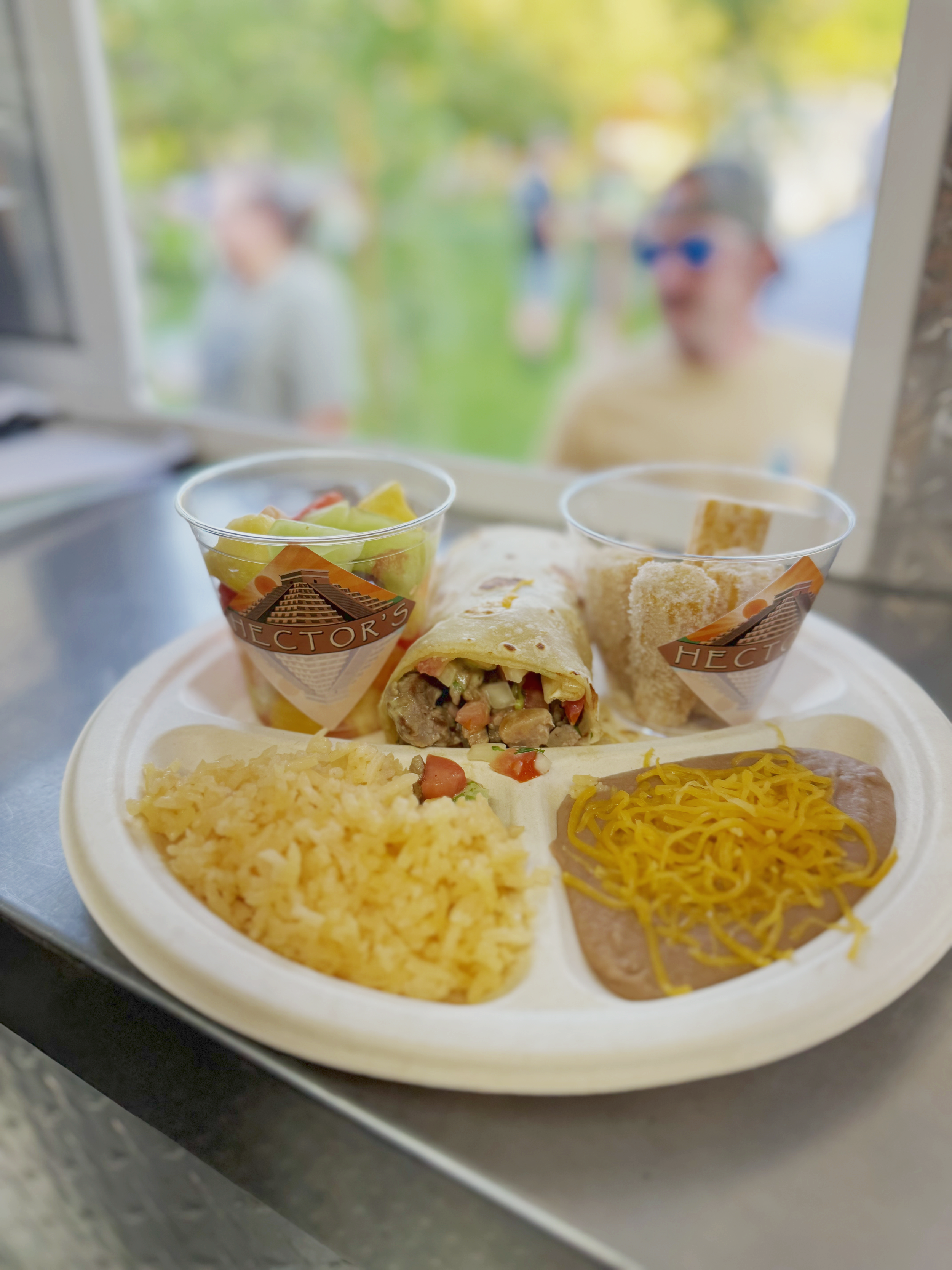 Tray with a burrito, rice, shredded cheese, beef and gravy, side salad, and a churro in a plastic cup, in front of a window with blurred people outside.