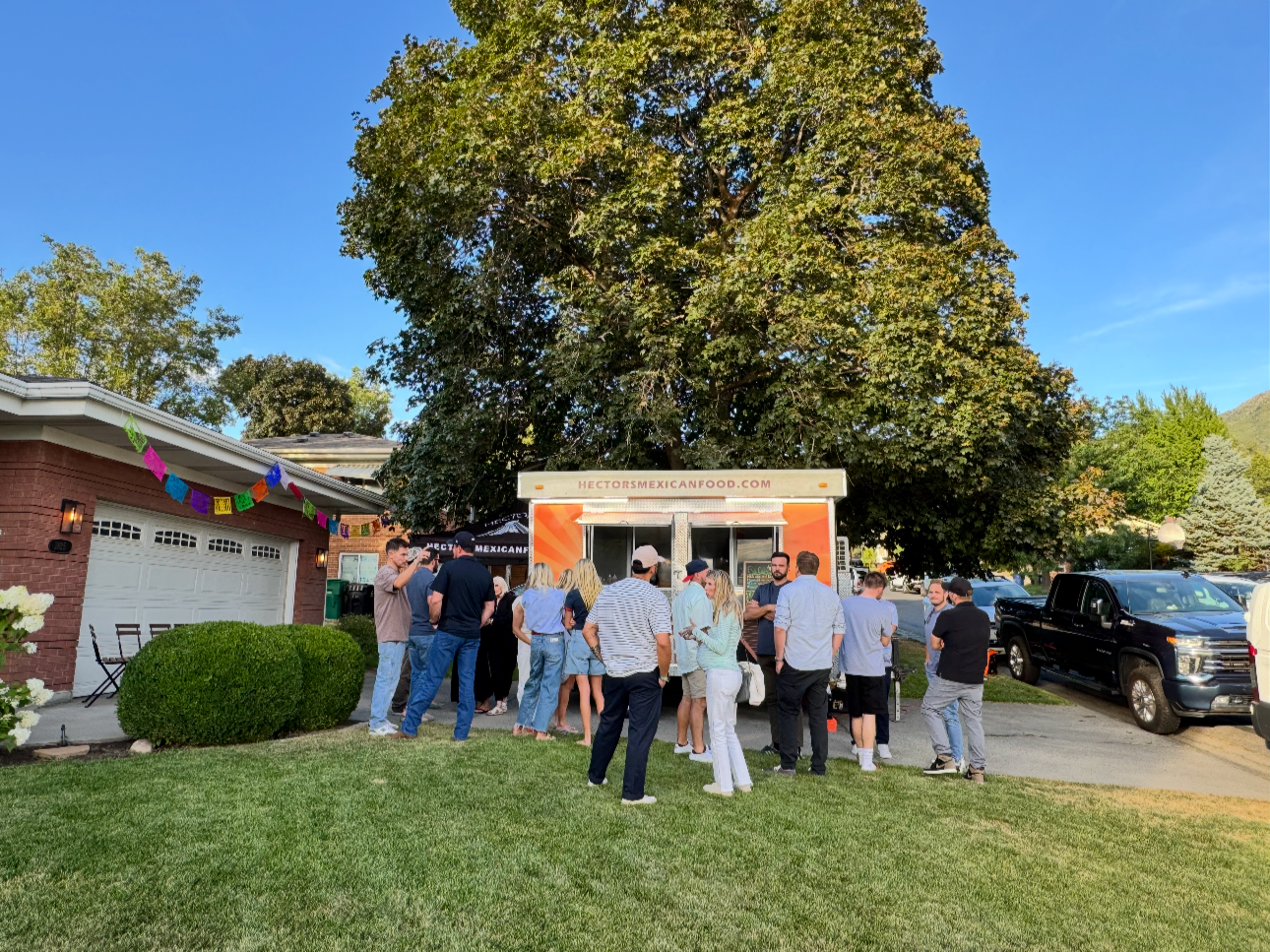 Group of people waiting in line at a food truck in a residential neighborhood with a large tree, blue sky, and parked cars.