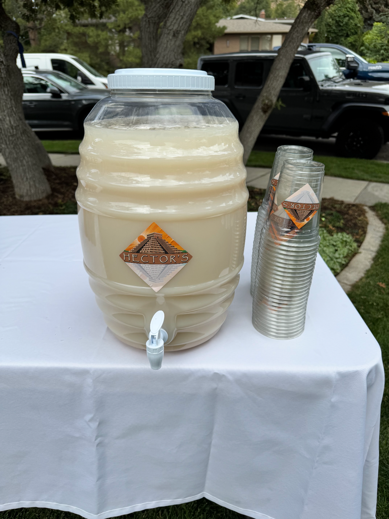 Large clear plastic water jug with a white lid and the Hector's logo, placed on a white tablecloth, with stacked clear plastic cups beside it, outdoors near trees and parked cars.