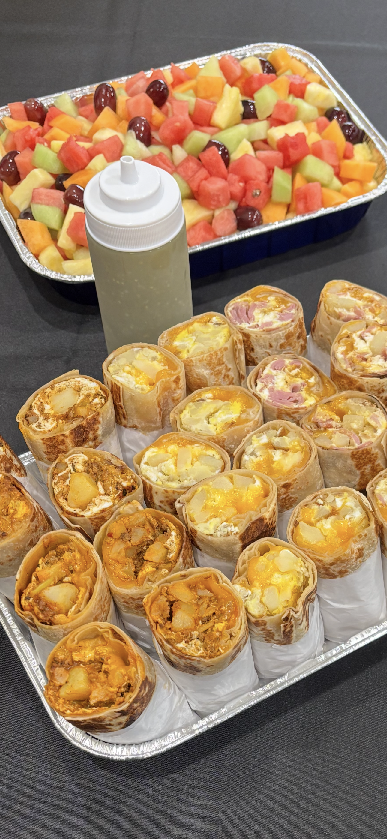 Tray of breakfast burritos with eggs, cheese, ham, and potatoes, alongside a bottle of sauces and a fruit salad with watermelon, cantaloupe, honeydew, pineapple, and grapes.