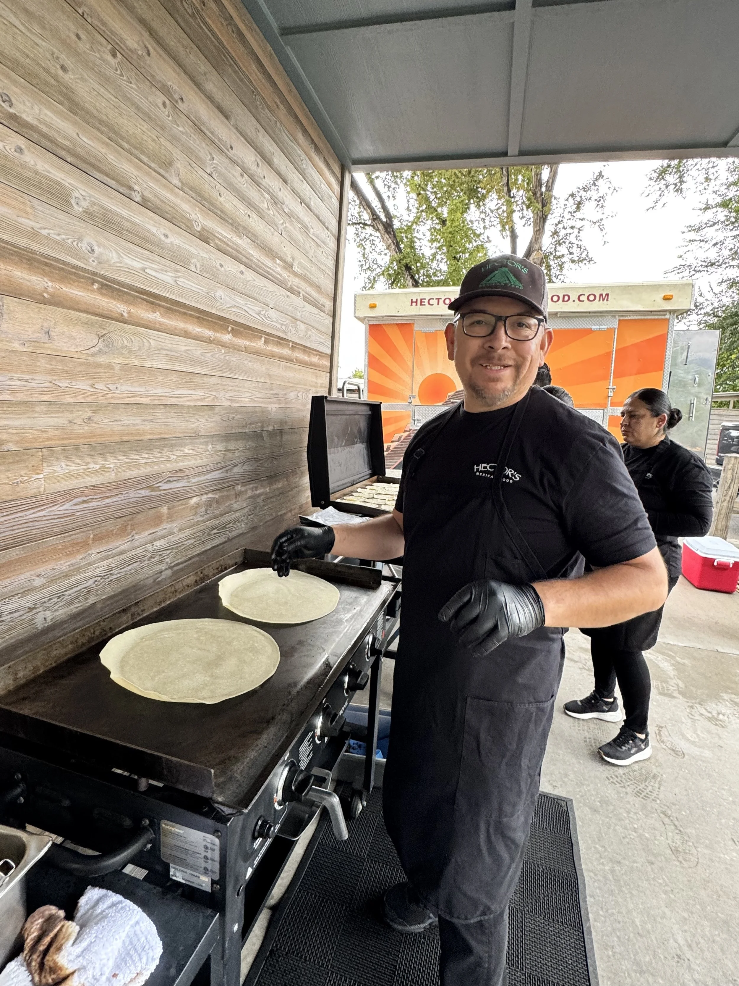 Man cooking two tortillas on a griddle at a food stand. He is wearing glasses, a black hat, a black apron, and black gloves. A woman is in the background and there is a food truck behind him.