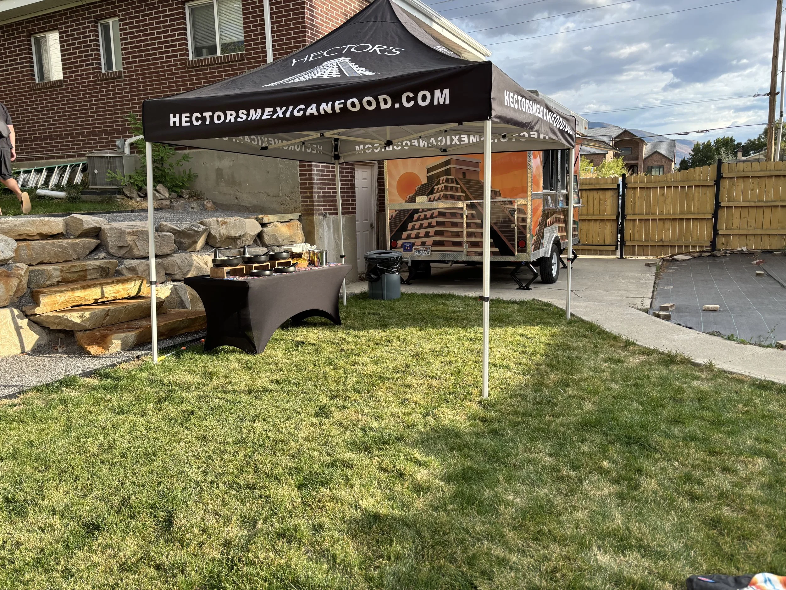 An outdoor food tent with the website 'hectormexicanfood.com' printed on it. There is a table with food and utensils underneath the tent, and a food trailer decorated with Mayan pyramid artwork behind it. The setting is a residential yard with a bric