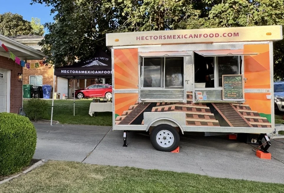 A food truck with a colorful exterior parked on a driveway in front of a house, with a table set up for outdoor dining nearby. The food truck has a website URL on top, and a chalkboard menu on its window.