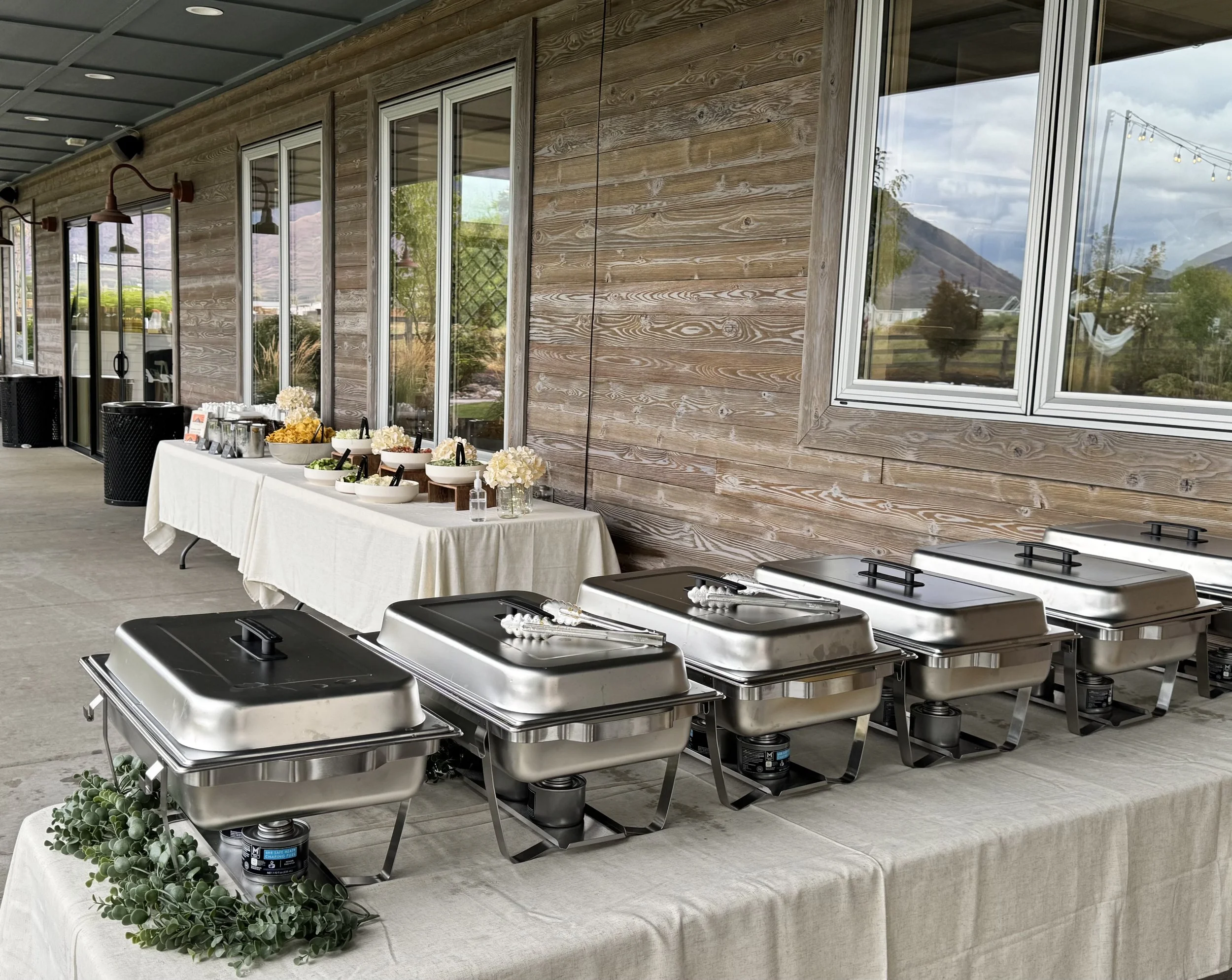 Buffet table with chafing dishes on a table with a beige tablecloth, set up outdoors against a wooden building with large windows, with mountains and trees visible in the background.