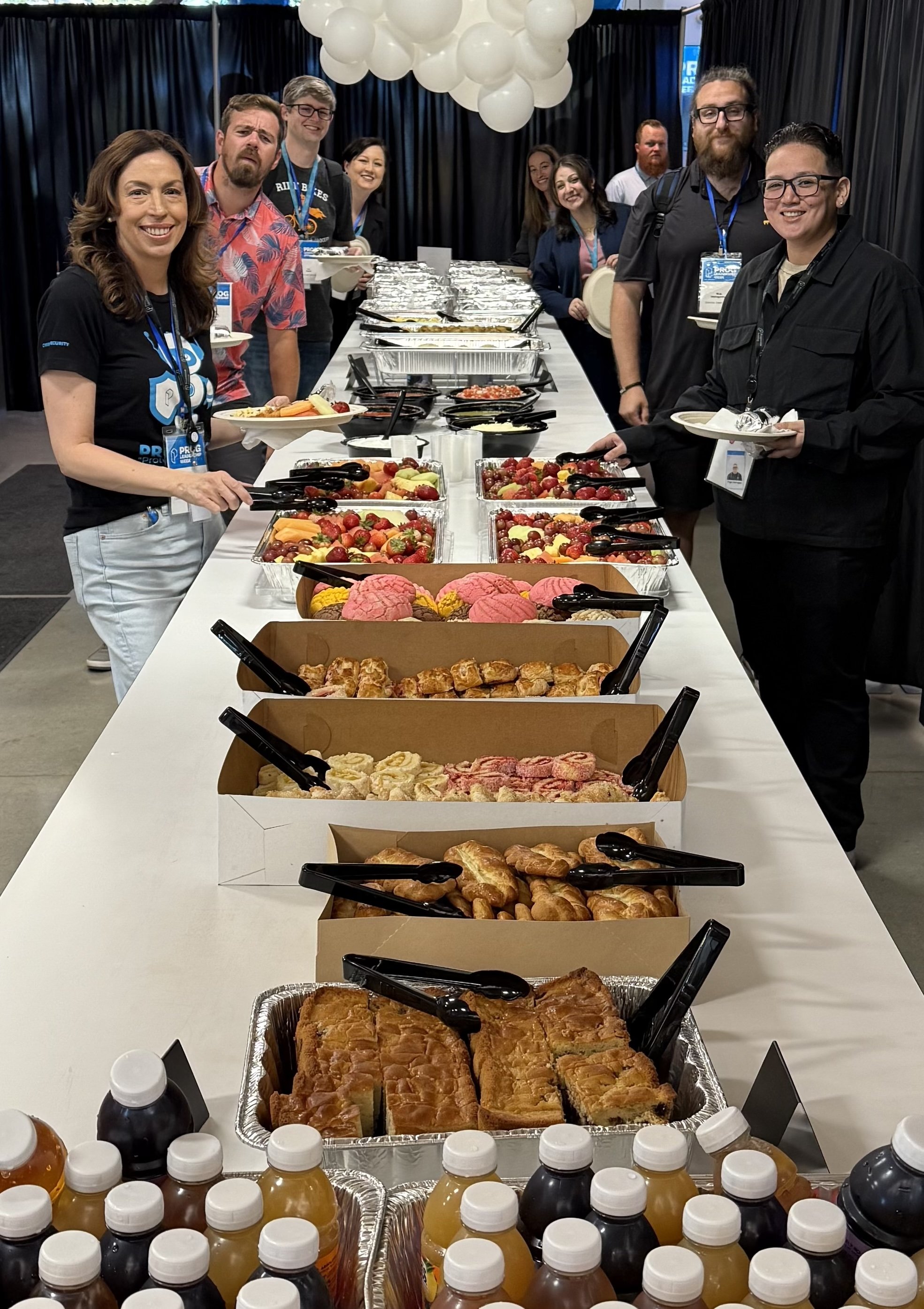 A group of people standing behind a long table filled with breakfast foods, including muffins, pastries, fruit, and bottled drinks, at a conference or event.