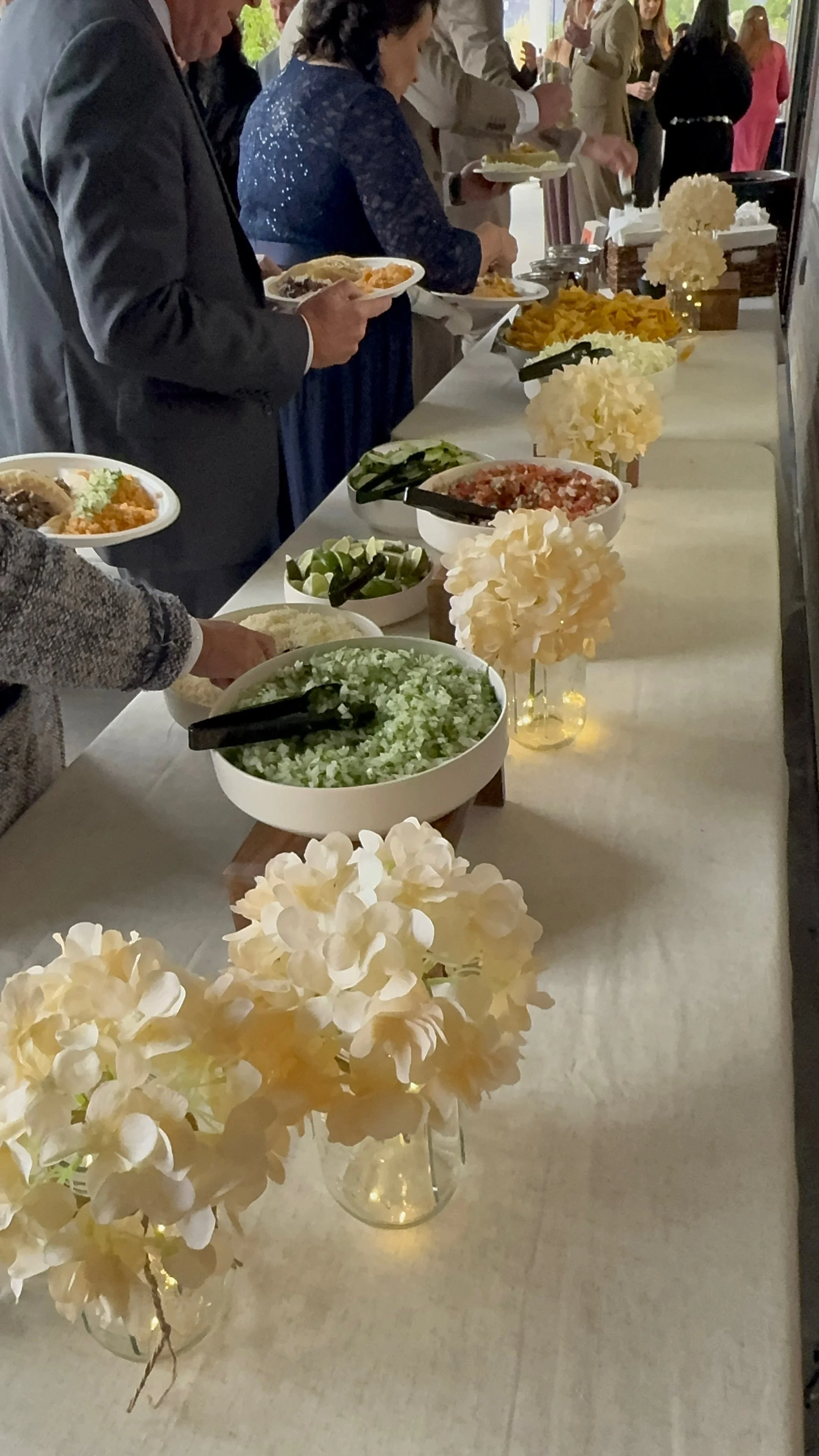 People serving themselves food at a buffet table decorated with white flowers in glass vases.