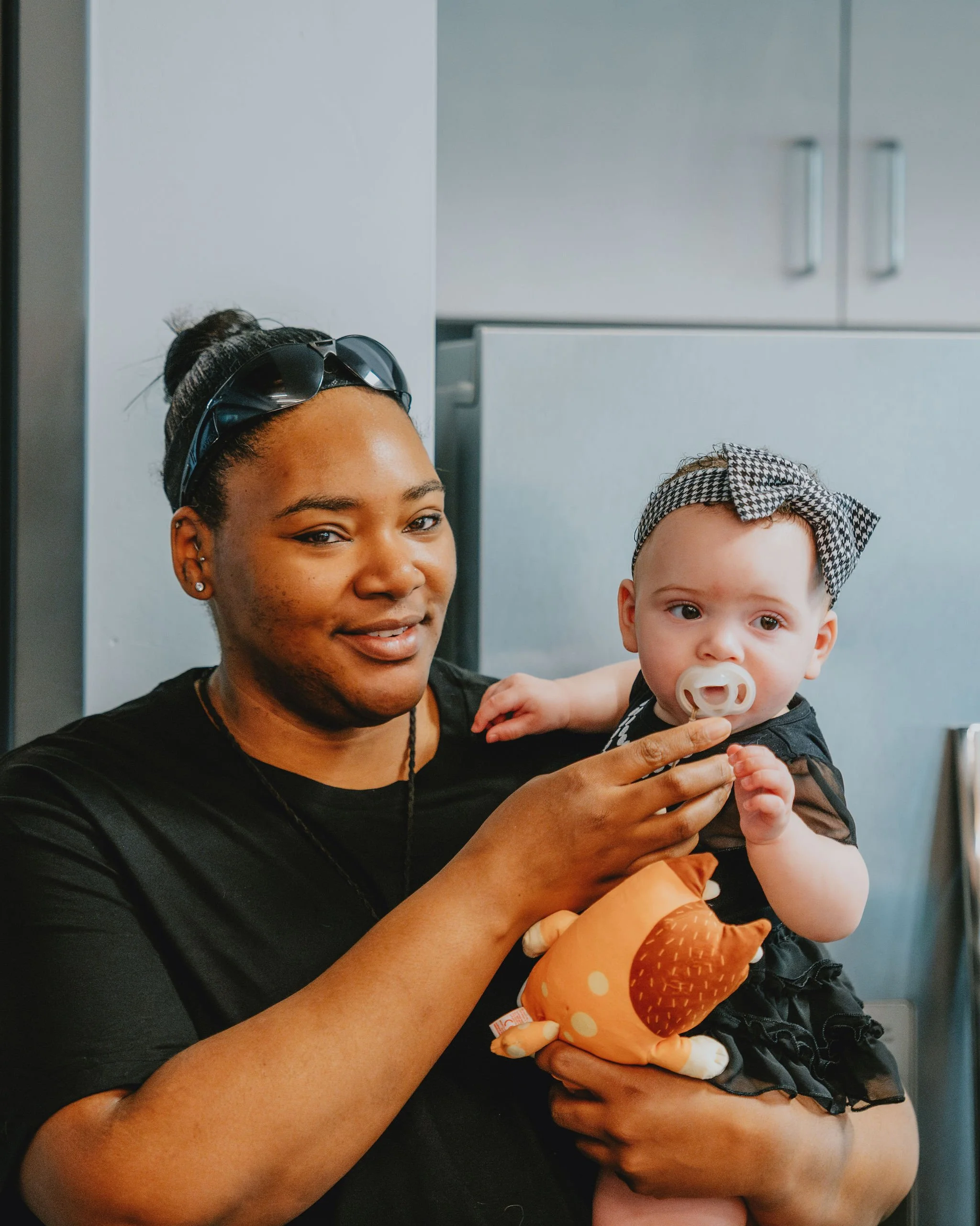 A woman holding a baby with a pacifier in her mouth. The woman is smiling, wearing a black top, and has sunglasses on her head. The baby has a headband with a bow and is holding a plush toy, looking curiously.