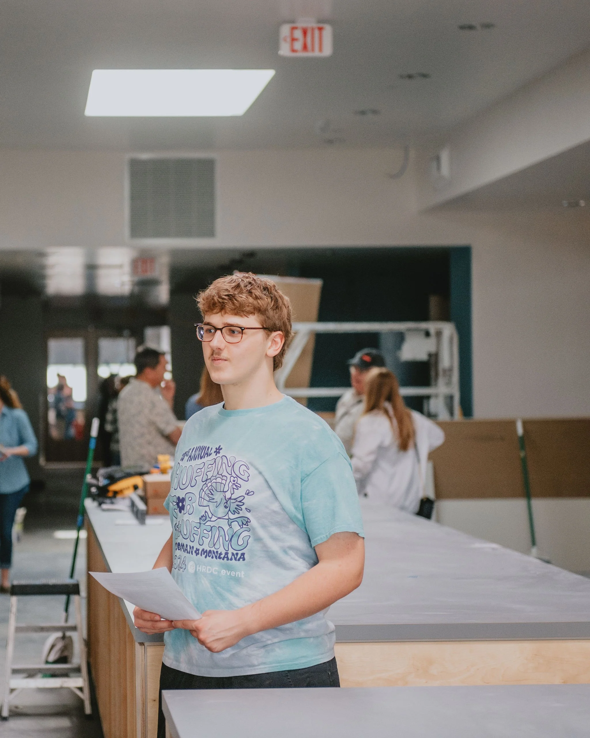 Young man with glasses holding papers inside a busy indoor space with people in the background and an illuminated exit sign on the ceiling.