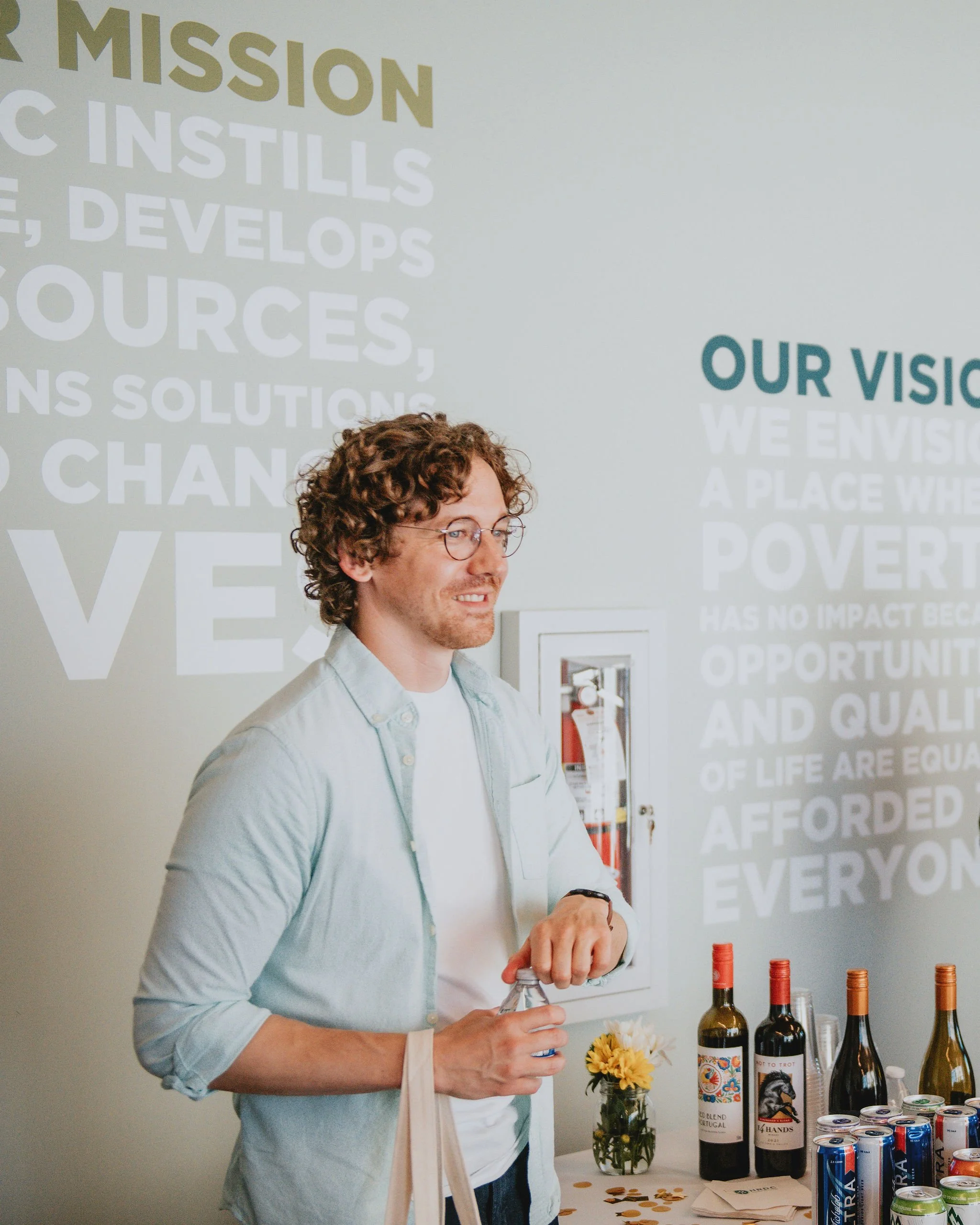 A man with curly hair and glasses standing next to a table with wine bottles and cans, in front of a wall with mission and vision statements.