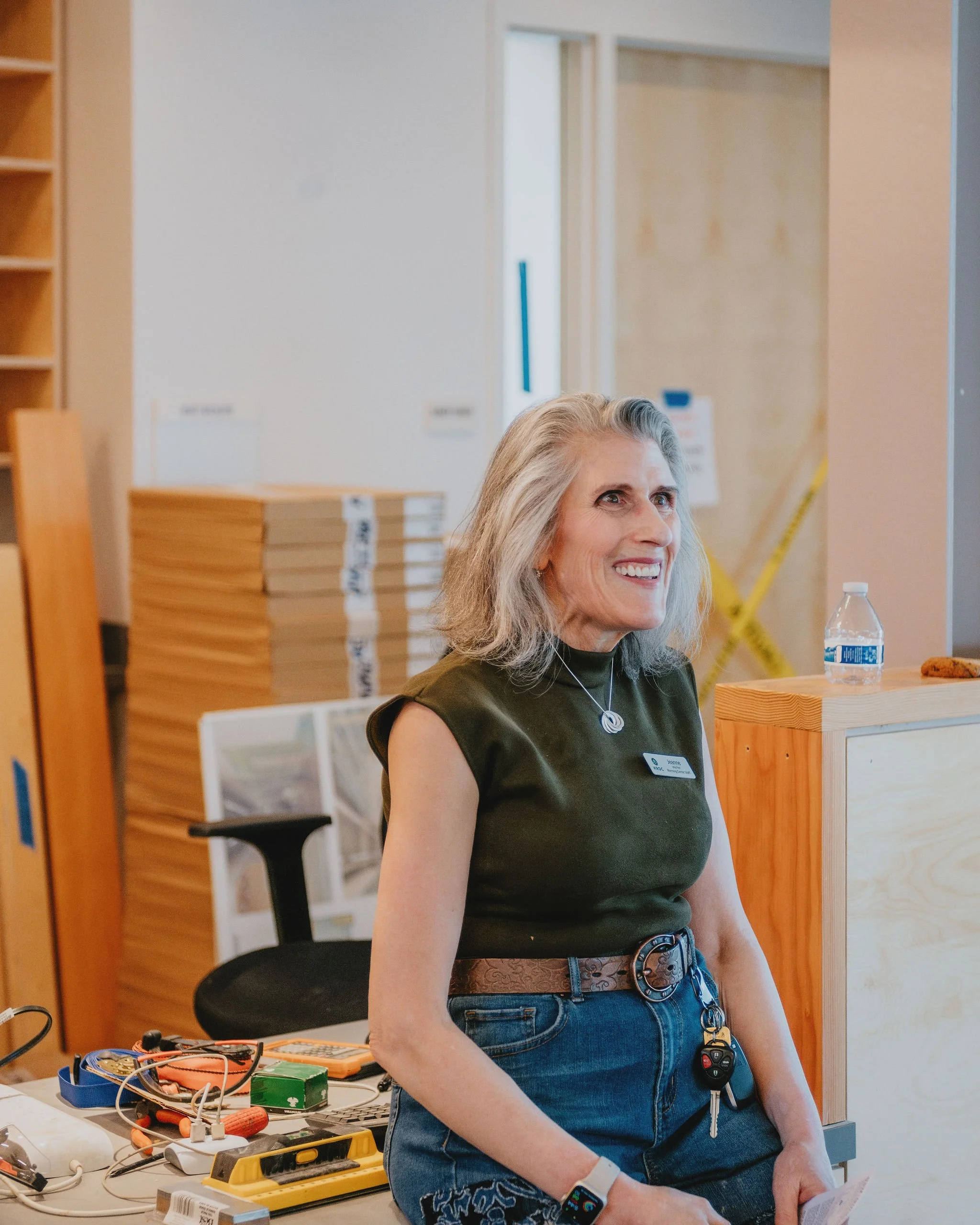 A smiling older woman with gray hair, wearing a dark green sleeveless top and jeans, standing behind a cluttered table in an office or workshop space.