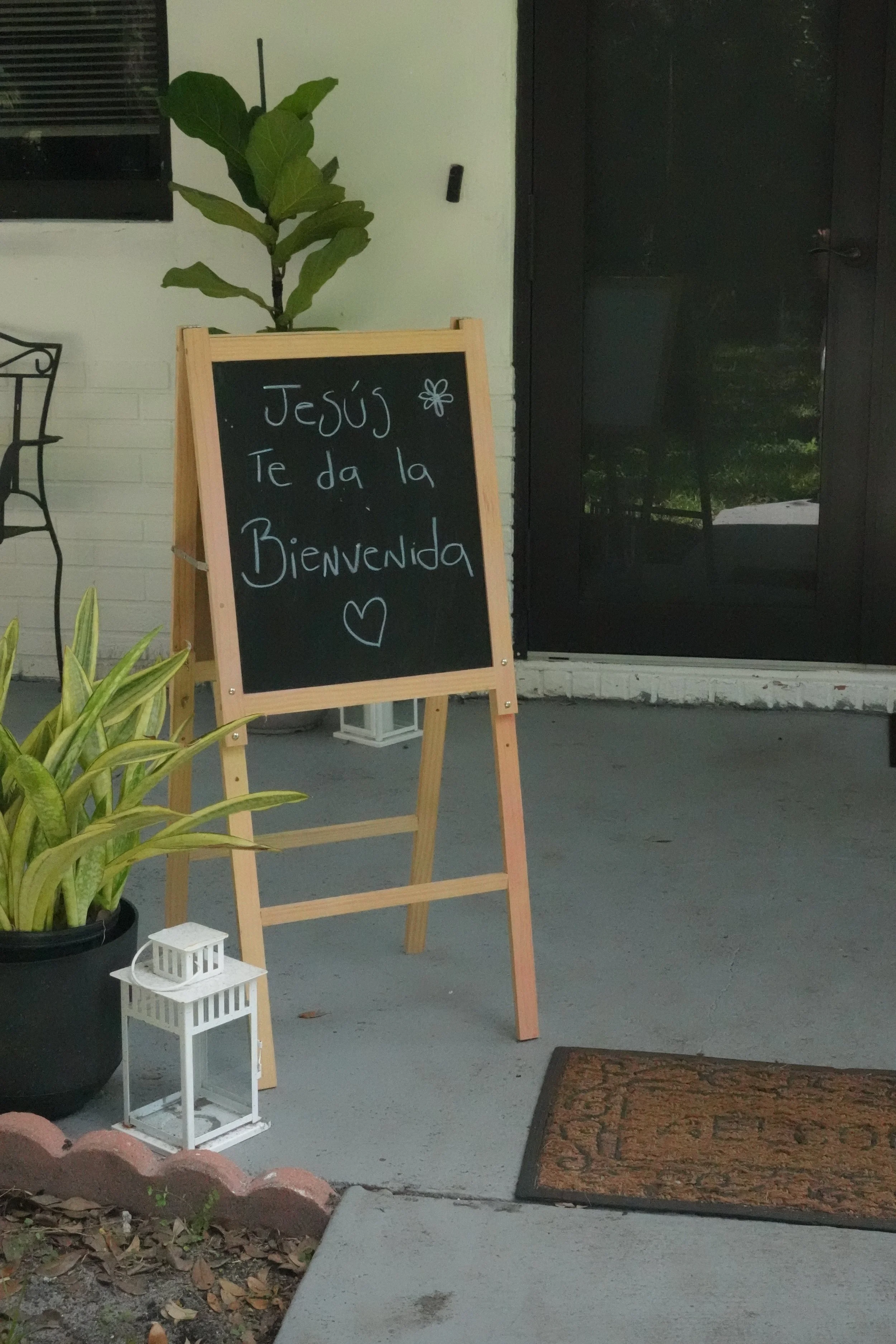 Un cartel en la entrada de una iglesia o establecimiento que dice 'Jesús te da la bienvenida' con un corazón dibujado debajo, acompañado de plantas y una pequeña lámpara blanca.