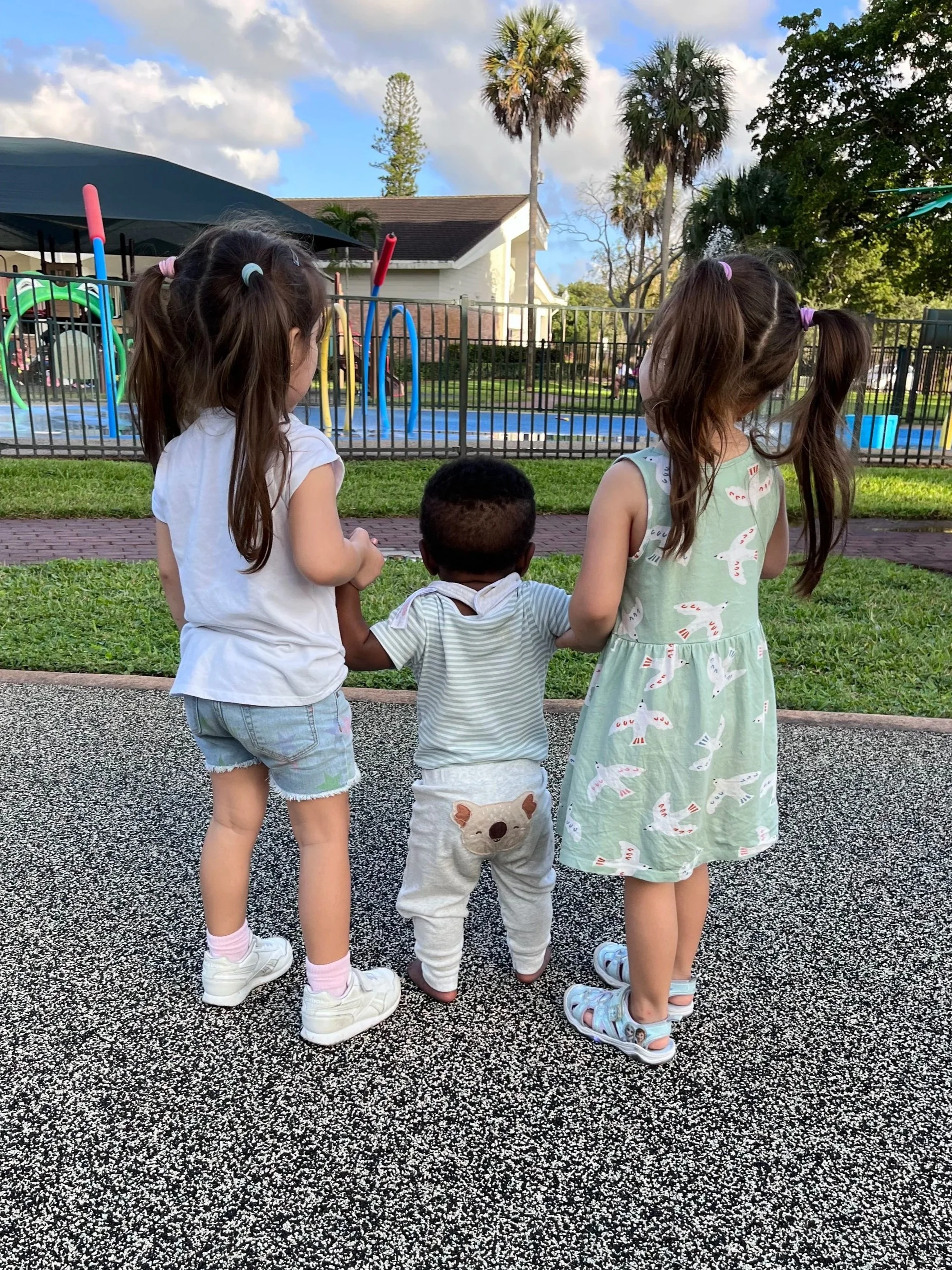 Tres niños pequeños de espaldas, sosteniéndose de las manos, en un parque con área de juegos y fuentes, con árboles y casas de fondo.