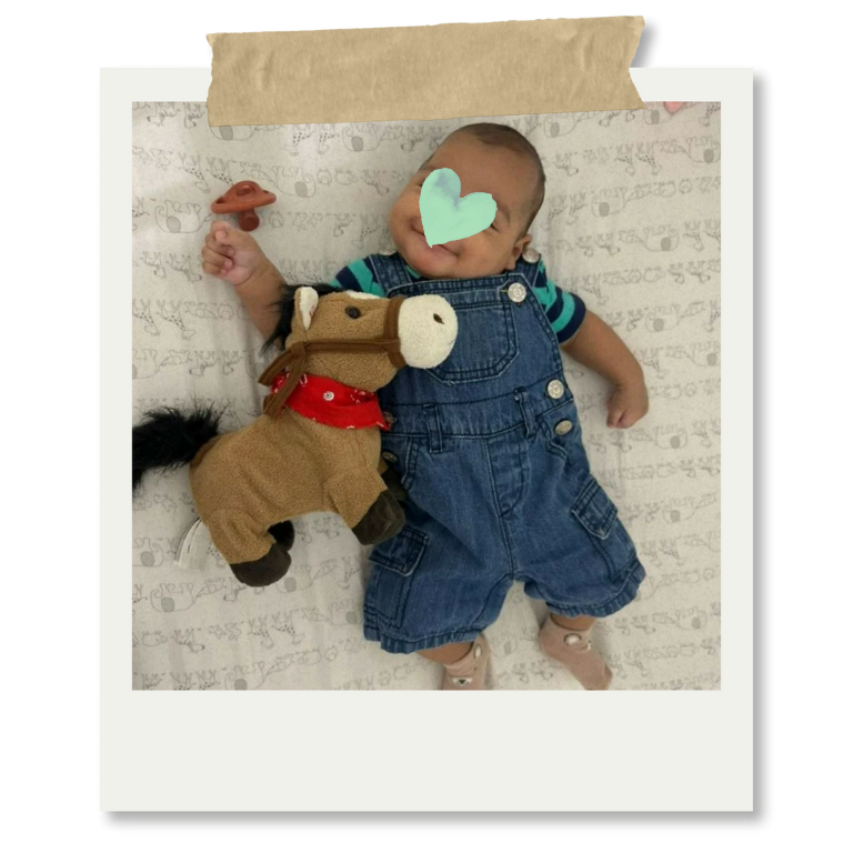 Bebé acostado en la cama con rostro cubierto por un corazón azul, sosteniendo un caballito de peluche y una chupeta, vestido con mameluco de mezclilla.