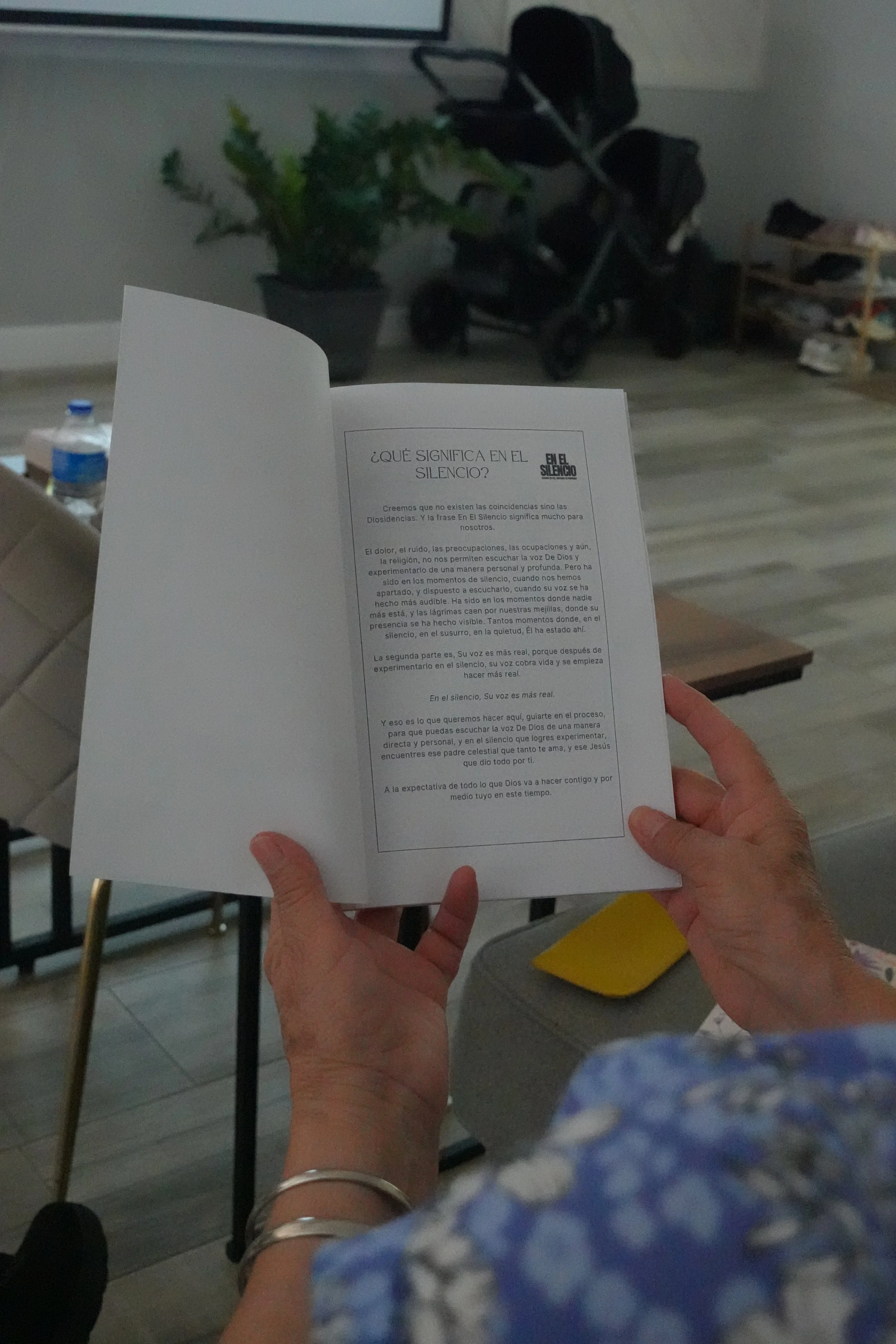 Persona sosteniendo un folleto con texto en español en un espacio interior, con una planta, una botella de agua, una silla y un carrito en el fondo.