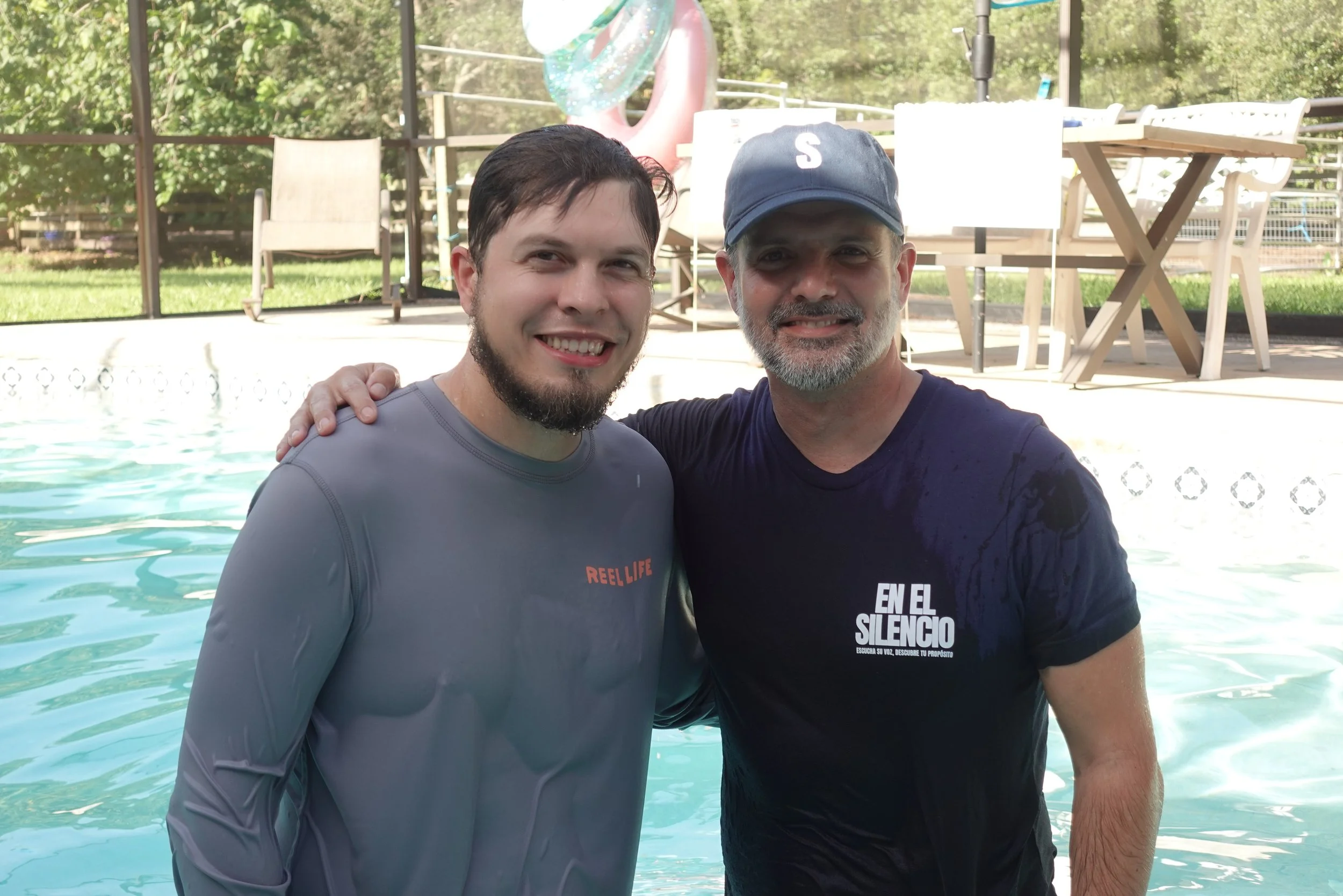 Dos hombres en la piscina, uno con camiseta gris y el otro con camiseta negra y gorra azul, sonriendo.
