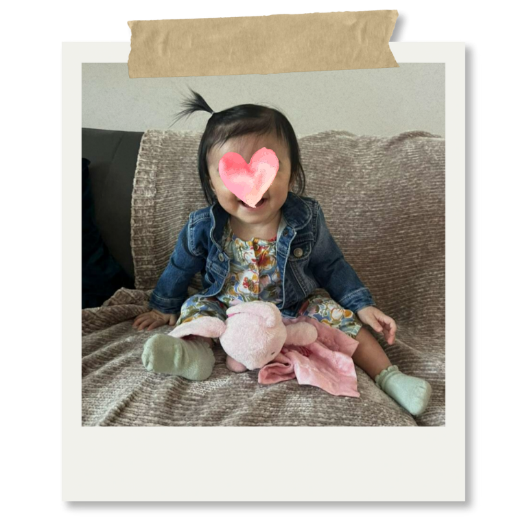 Niña pequeña sentada en un sofá, sonriendo, con la cara cubierta por un corazón rosa, sosteniendo un peluche de conejo en las piernas, usando una chaqueta de mezclilla y un vestido colorido.