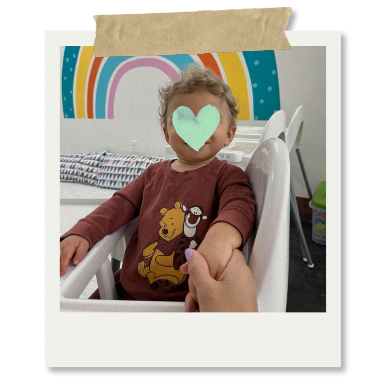 Niño sentado en una silla de madera, con rostro cubierto con un corazón de color verde, rodeado de un decorativo mural con arcoíris en la pared de fondo y accesorios infantiles en un ambiente interior.