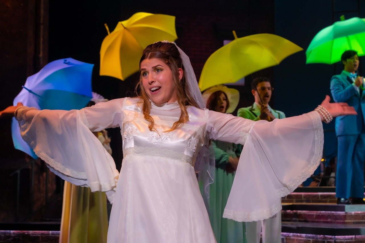A woman in a white dress singing on stage with colorful umbrellas held by others behind her.