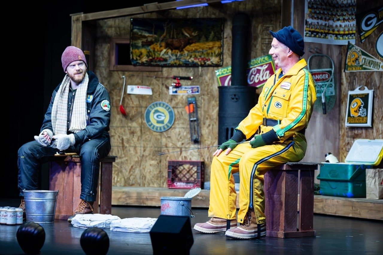 Two men sitting on wooden boxes in a rustic, decorated room. One man is dressed in a yellow jacket and pants with green accents, and the other wears a pink beanie, scarf, gloves, and casual clothing. They are engaged in a conversation, surrounded by