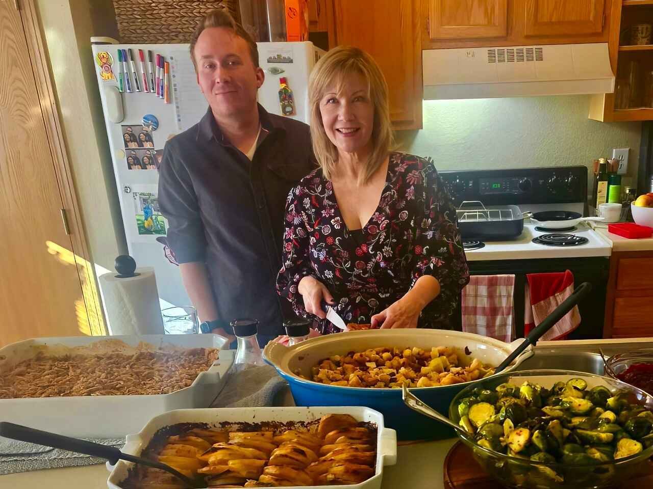 Two people in a kitchen with a variety of dishes prepared, including roasted Brussels sprouts, a casserole, baked apples, and a dish with breadcrumbs. There is also a stove with hanging towels and a refrigerator with photos and magnets.