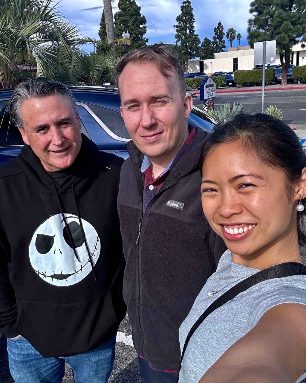 Three people smiling outdoors with palm trees and a blue car in the background.