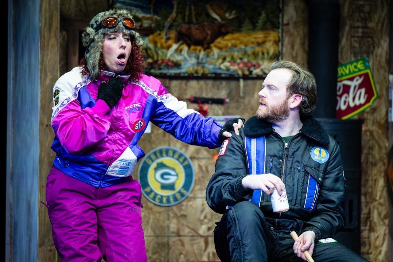 A woman in a colorful racing jacket and fur-trimmed hat appears surprised or shocked as she touches her throat, while a man with a drink in his hand looks at her with concern. Behind them, there is a wall with a yellow and blue logo and a vintage Coc