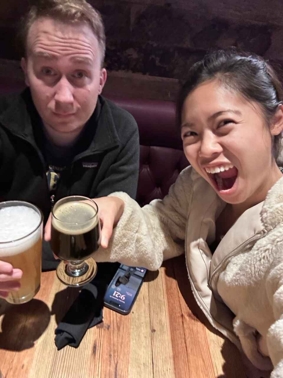 Two people sitting at a wooden table, smiling and holding drinks—a glass of beer and a dark beverage. A phone is placed on the table showing the time.