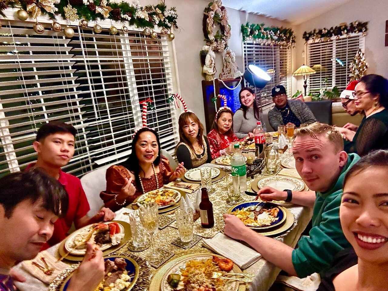 A group of people celebrating around a Christmas dinner table decorated festively with greenery and lights, sharing a meal. They appear happy and engaged, with plates of food and beverages on the table. The room is adorned with holiday decorations.