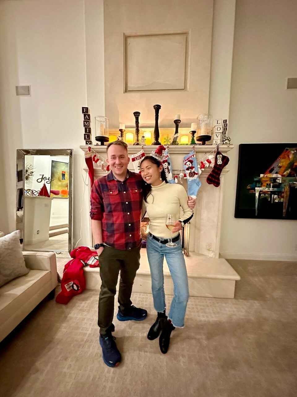 Two people smiling in front of a decorated fireplace with Christmas stockings, candles, and festive decorations.
