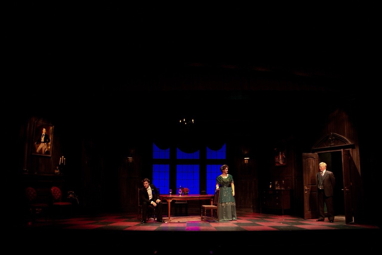 Stage scene with three actors in period costume, set in a dark, elegant room with large blue window, portrait, and wooden furniture.