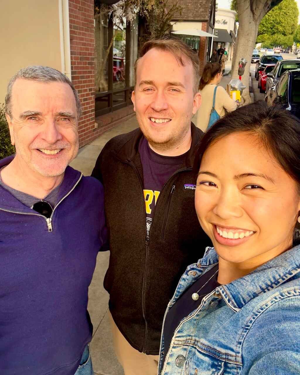 Three people taking a selfie on a sidewalk, smiling and standing close together, with a brick building and trees in the background.