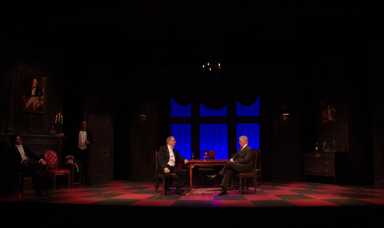 A theatrical stage set resembling a vintage room, with four men dressed in tuxedos. Two men sit at a table in the center, facing each other, with glasses of wine in front of them. Two men stand to the sides, one seated on a red chair and the other st