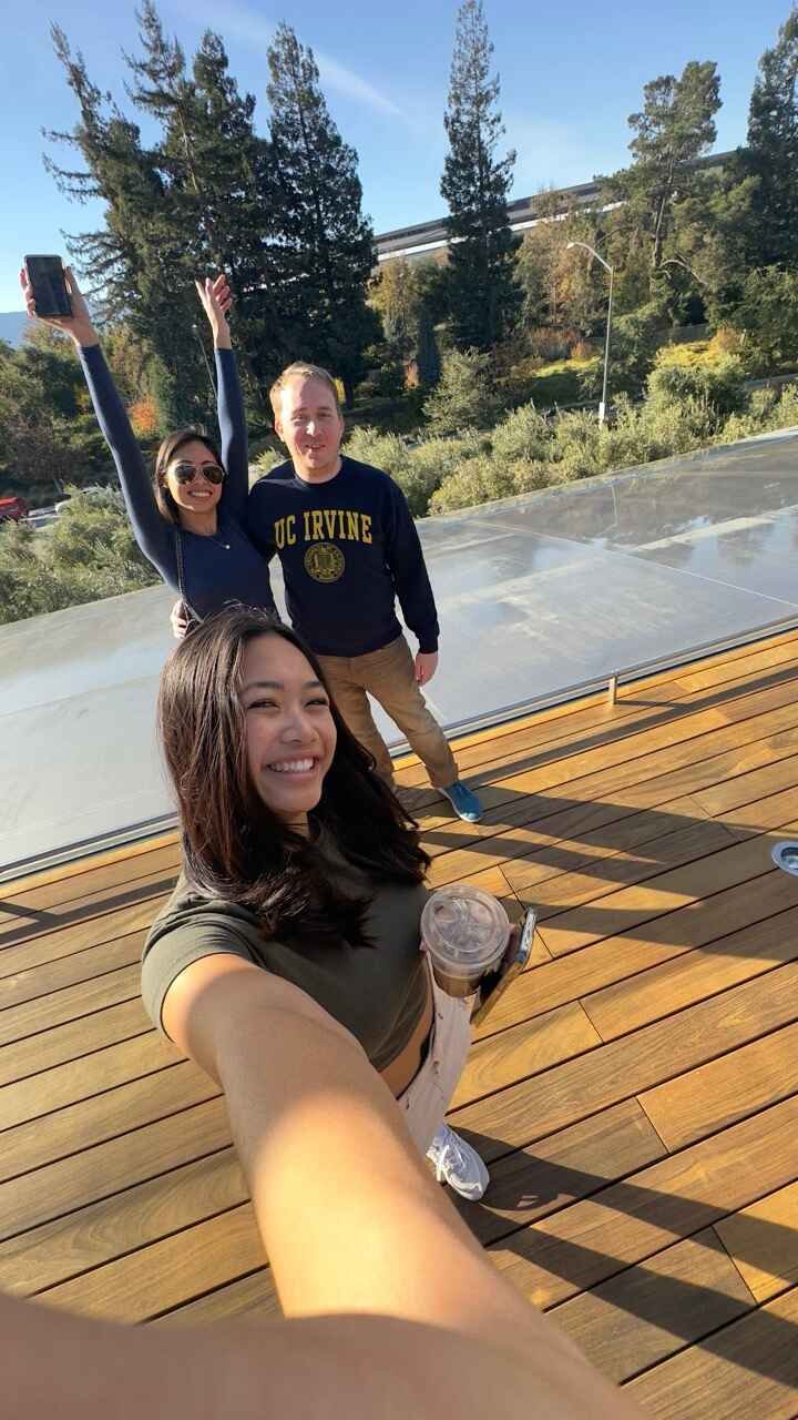 Three people taking a selfie on a wooden deck with trees in the background. One person holds a drink and smiles at the camera. Another is raising hands in excitement, wearing sunglasses. The third person wears a UC Irvine sweatshirt.