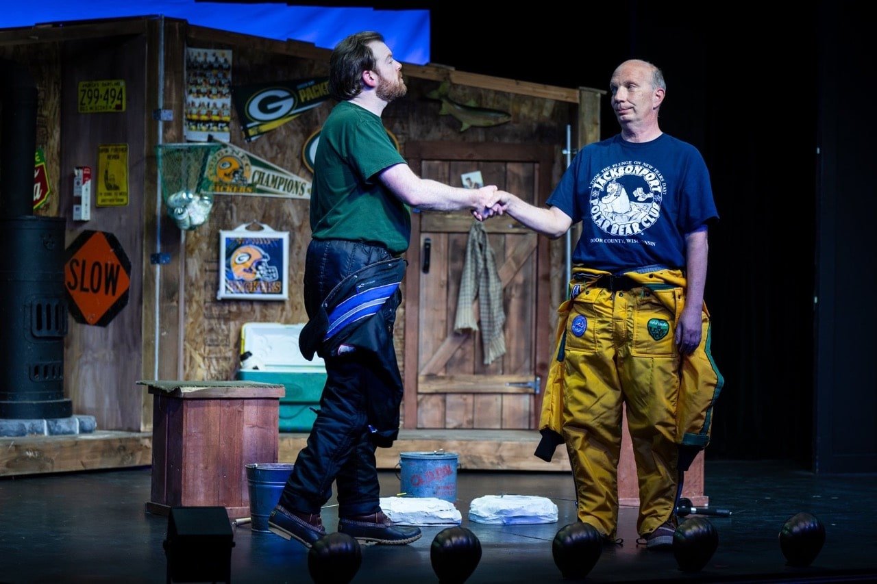 Two men shaking hands on stage in front of a wooden backdrop decorated with sports memorabilia, signs, and a door. One man has long hair and is wearing a green shirt, while the other has short hair and is wearing a blue shirt and yellow pants with pa