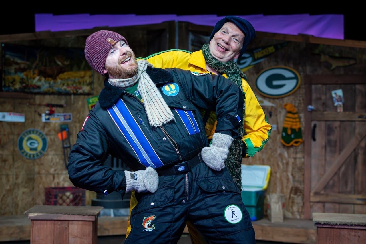 Two men smiling and posing playfully indoors, with wooden walls and sports team logos in the background.