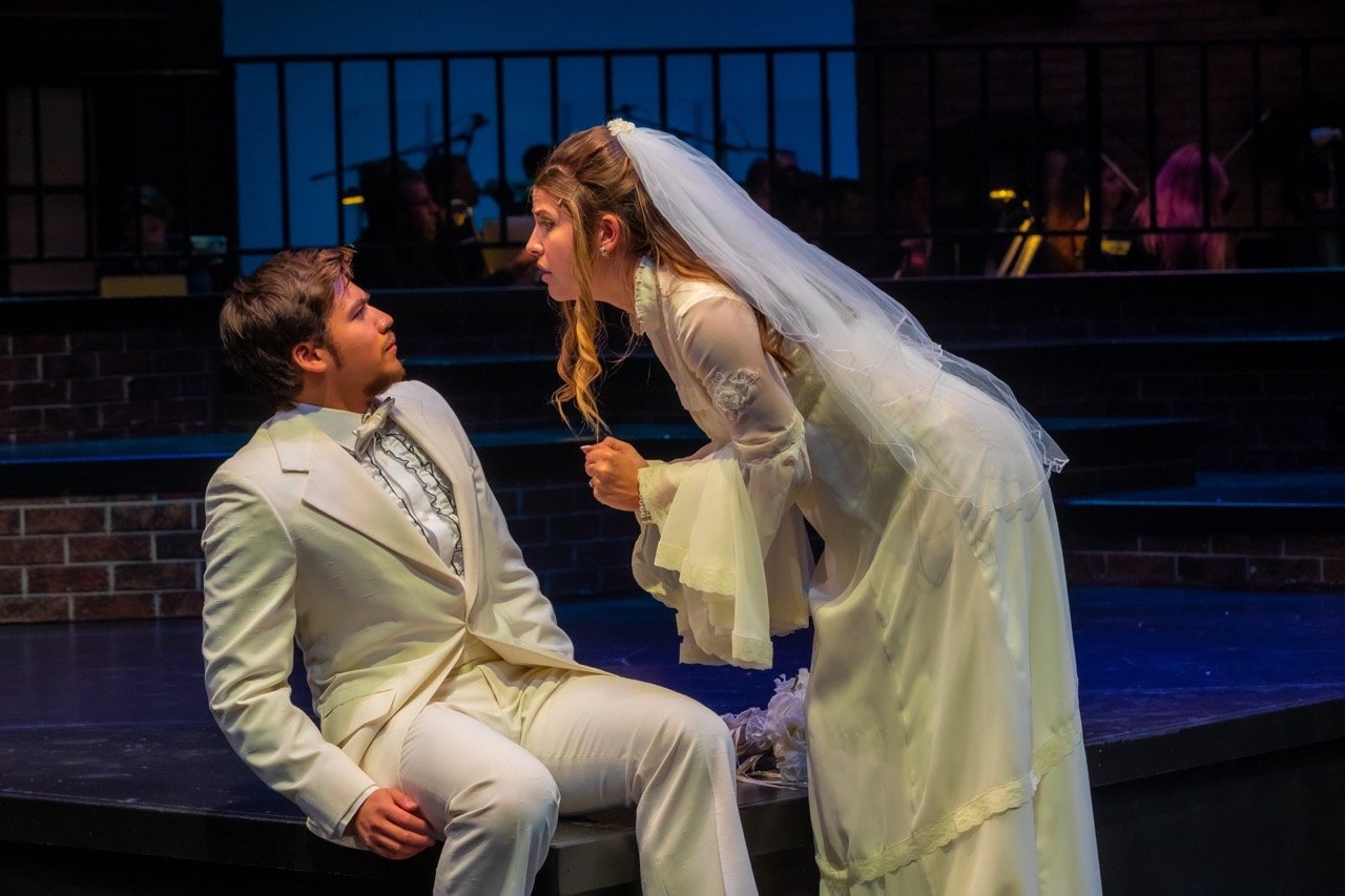 A woman in a wedding dress leaning forward and talking to a seated man in a white tuxedo on a stage.