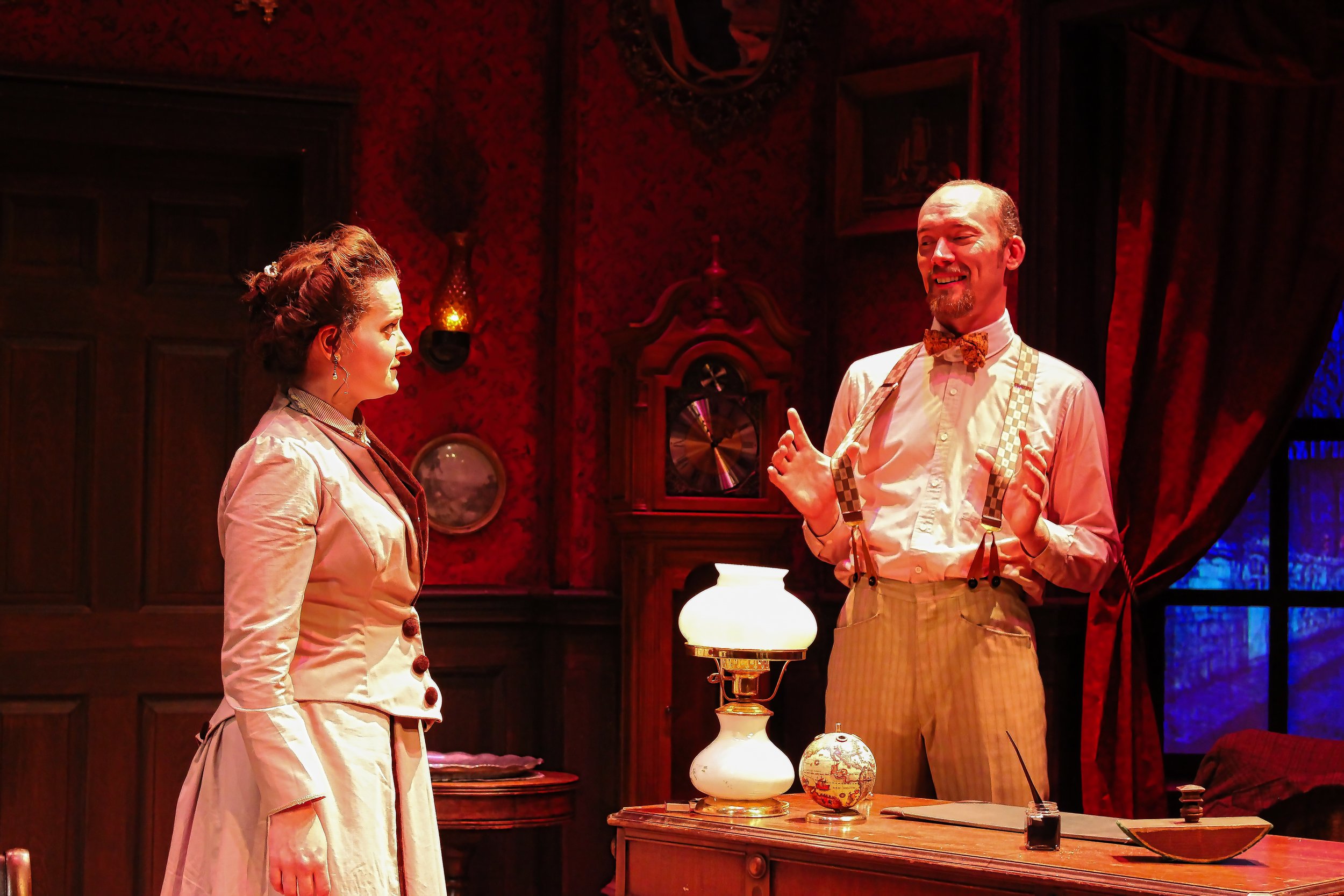Two characters, a woman and a man, having a conversation in a vintage-style room with red wallpaper, a clock, and a lamp on the desk.
