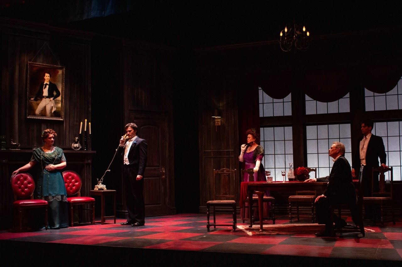 Actors performing in a theatrical play set in a vintage-style room with dark wood paneling, red checkered floor, and large windows in the background.